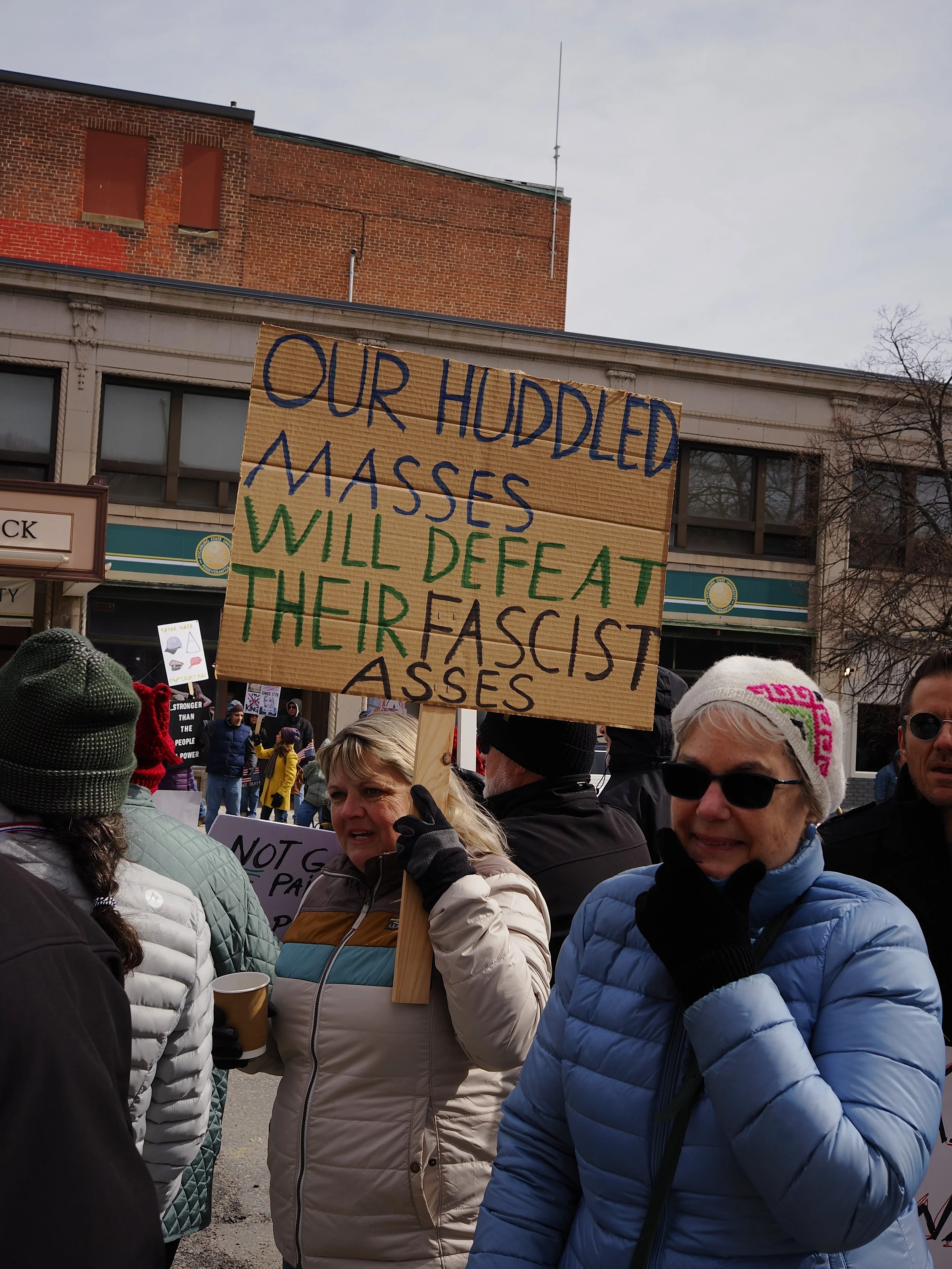 Woman holding sign that says "Our Huddled Masses Will Defeat Their Fascist Asses" at No Kings III in Fitchburg by Mass 50501.