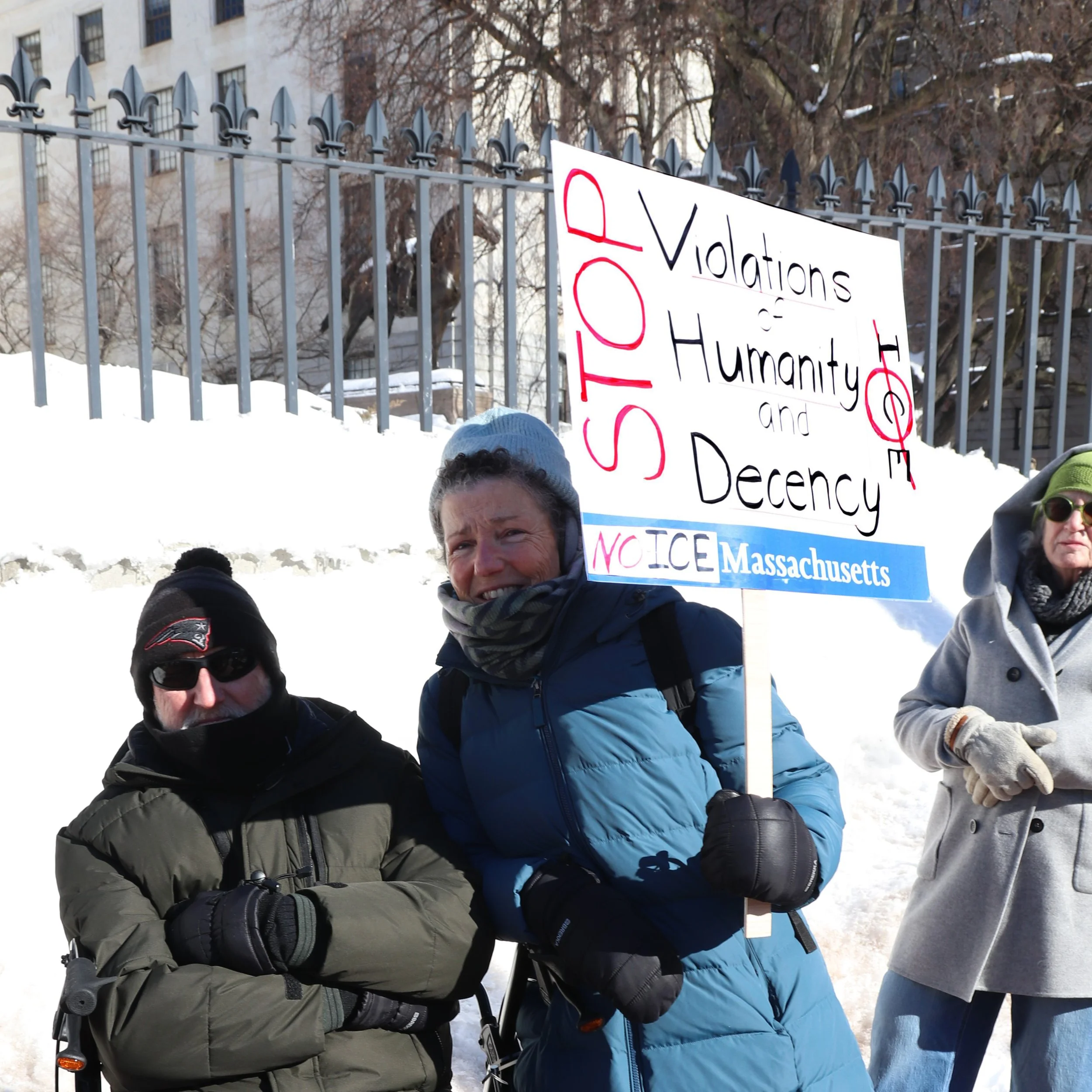 A photograph of a woman holding a sign saying “Stop violations of humanity and decency”, “No ICE Massachusetts”, and a crossed-out “ICE”, with a man seated next to the woman and another protester behind them.