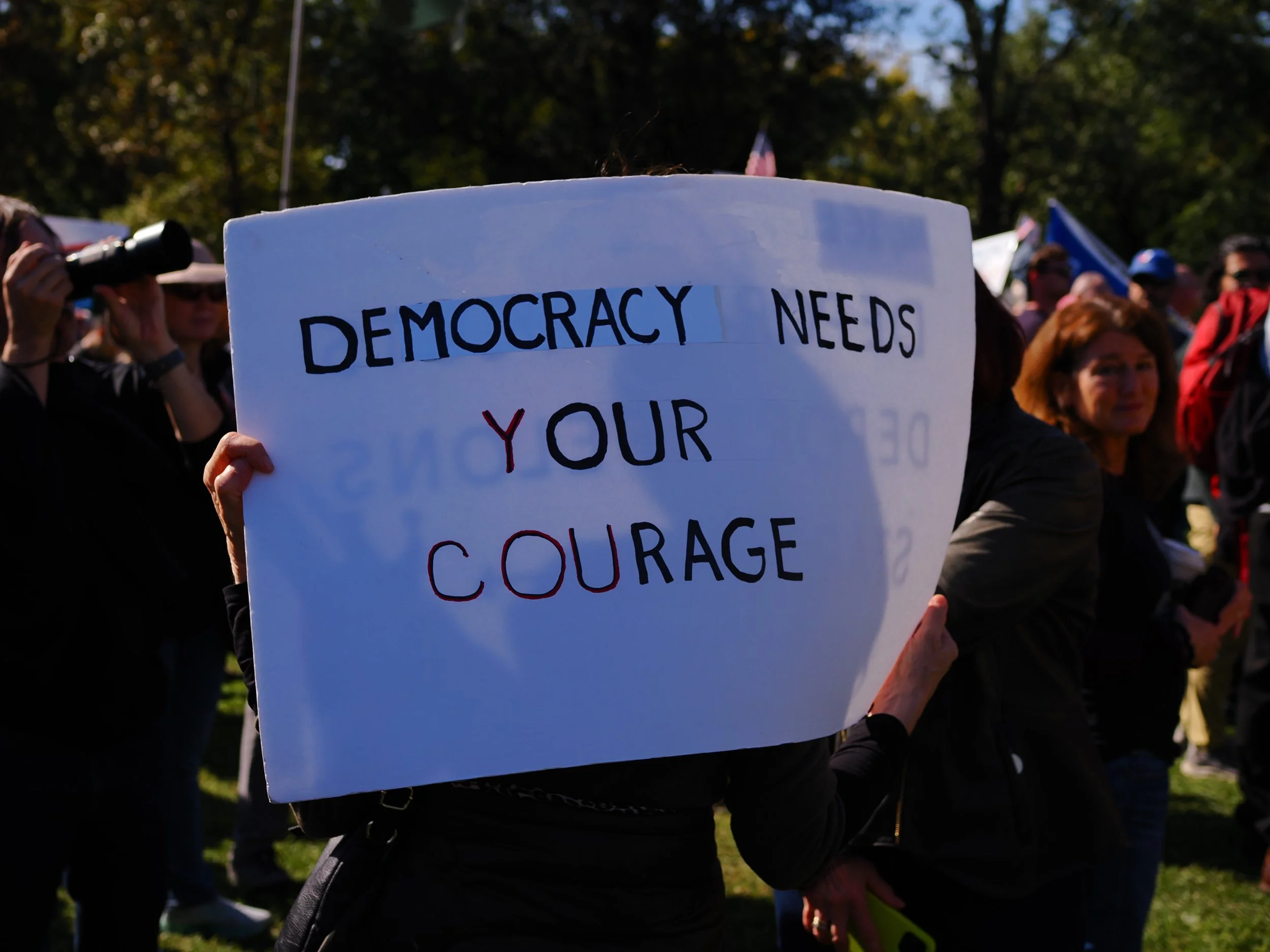 A sign saying “Democracy needs your courage” with a crowd of people in the background.