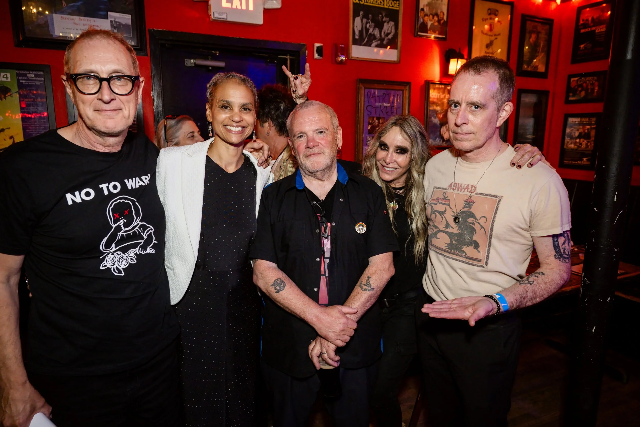 Five people standing in a row in the Burren, a bar and performing-arts venue in Somerville, Massachusetts, with someone doing the “rock on” sign with their hand from behind them.