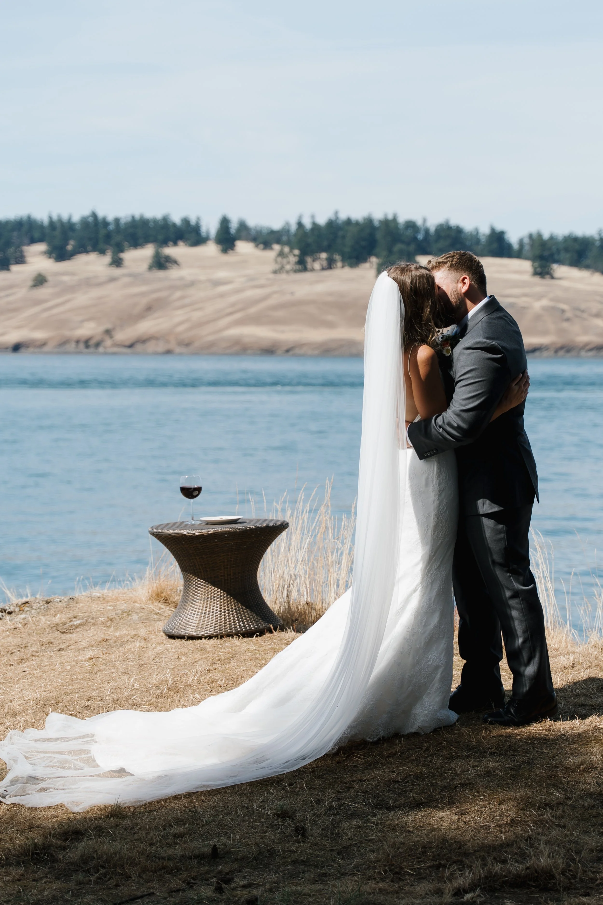 A bride and groom kissing near a lake, with a small table holding a glass of red wine nearby.