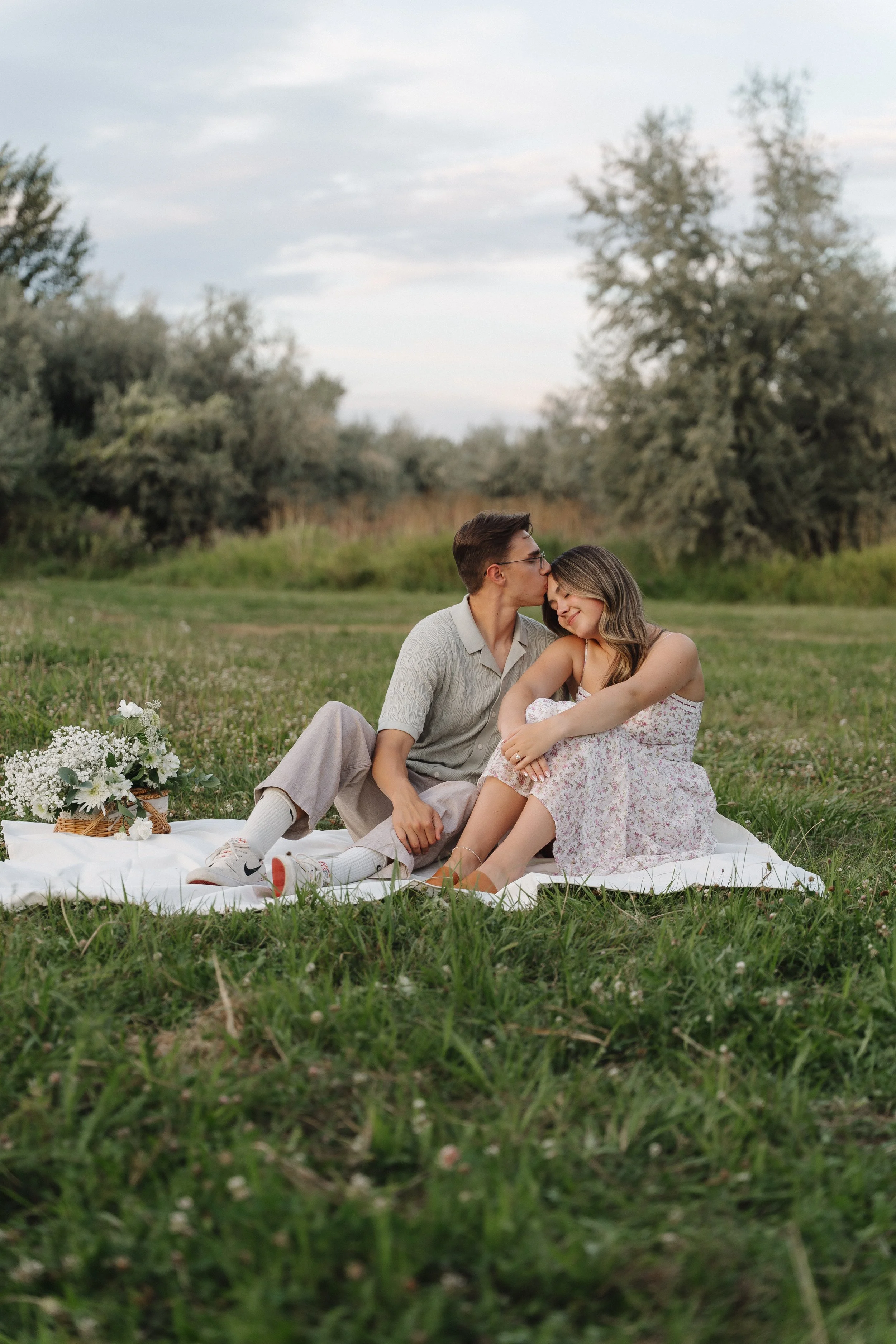 A couple sitting on a white blanket in a park, with the man kissing the woman's forehead, surrounded by greenery and trees.