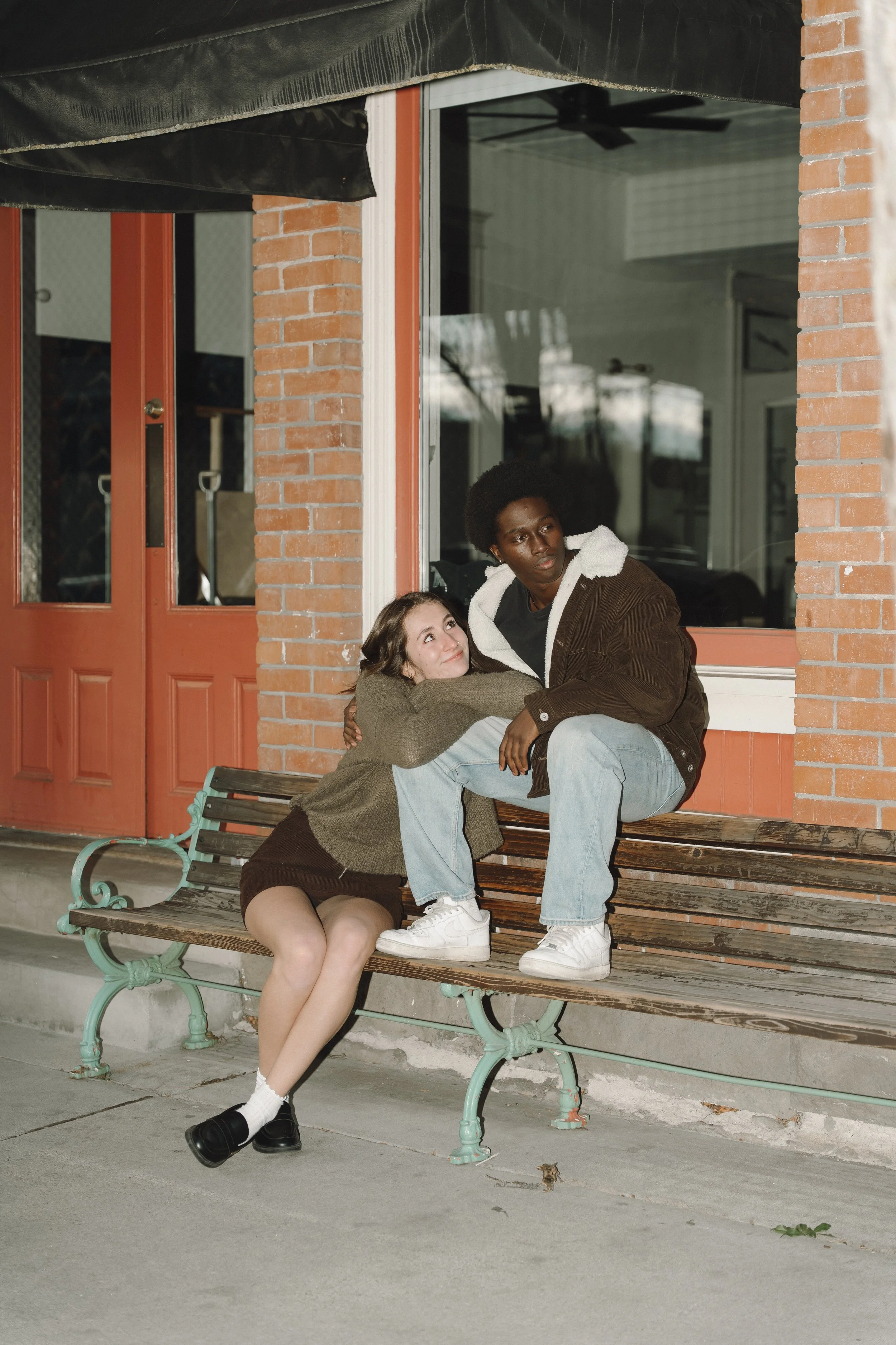 A young woman and a young man sitting on a wooden bench outside a building with red brick and large window. The woman is resting her head on the man's lap, and they appear to be having a heartfelt conversation.