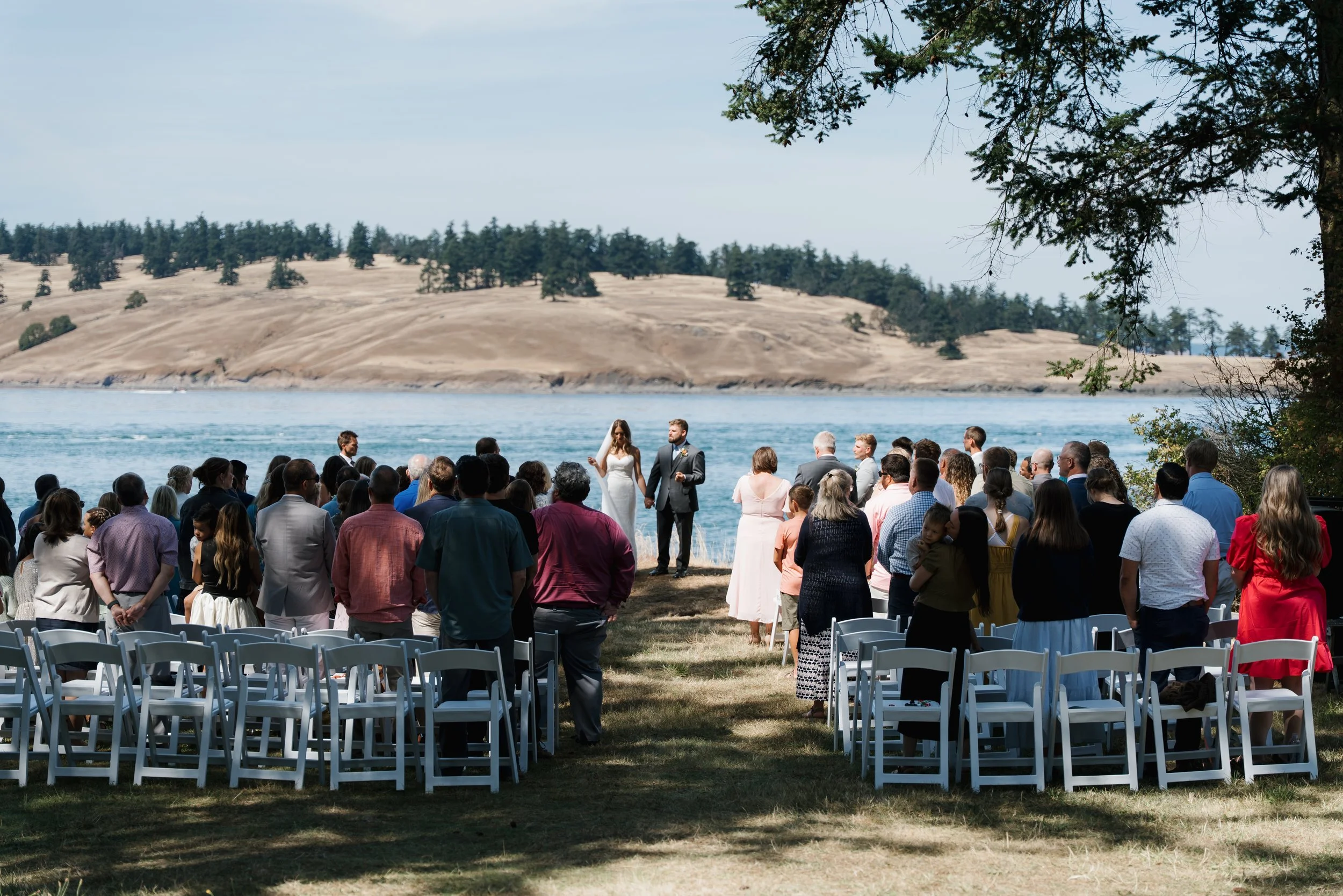 Outdoor wedding ceremony near a lake with guests standing and sitting, a bride and groom at the altar, tree branches overhead, and rolling hills in the background.