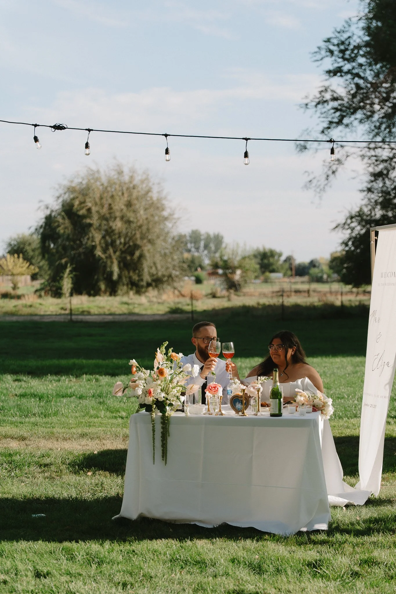 A couple at a wedding reception raising glasses of wine at a decorated outdoor table with flowers, a bottle of champagne, and a framed photo, set on a grassy field with trees and string lights overhead.