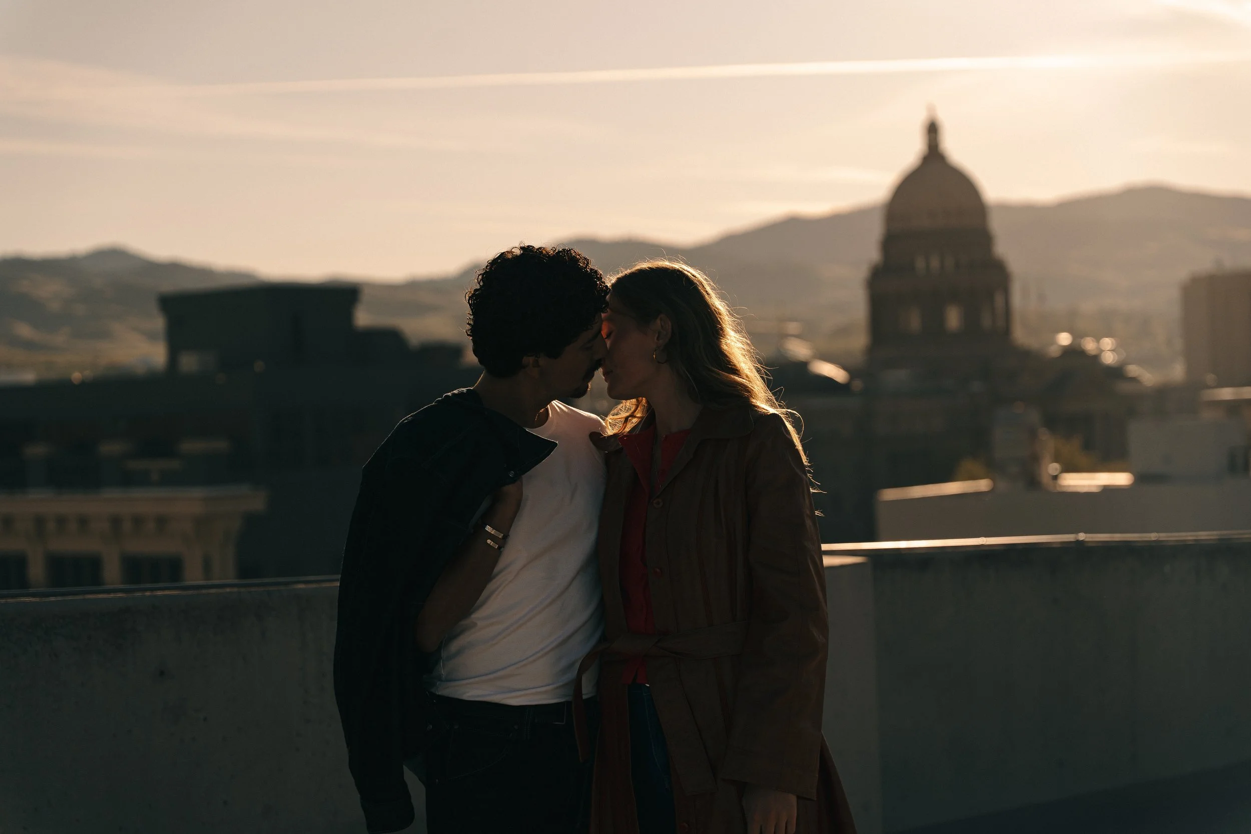 A couple sharing a kiss at sunset on a rooftop in San Francisco, with city buildings and the Capitol building in the background.