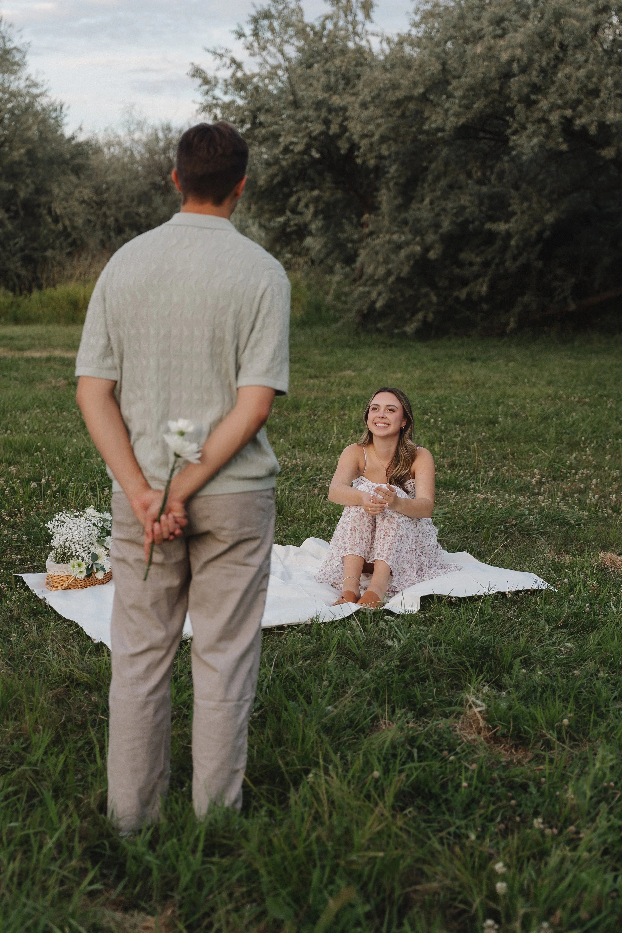 A couple sharing a romantic moment outdoors, with the woman sitting on a blanket and smiling at the man standing before her in a grassy field with trees in the background.
