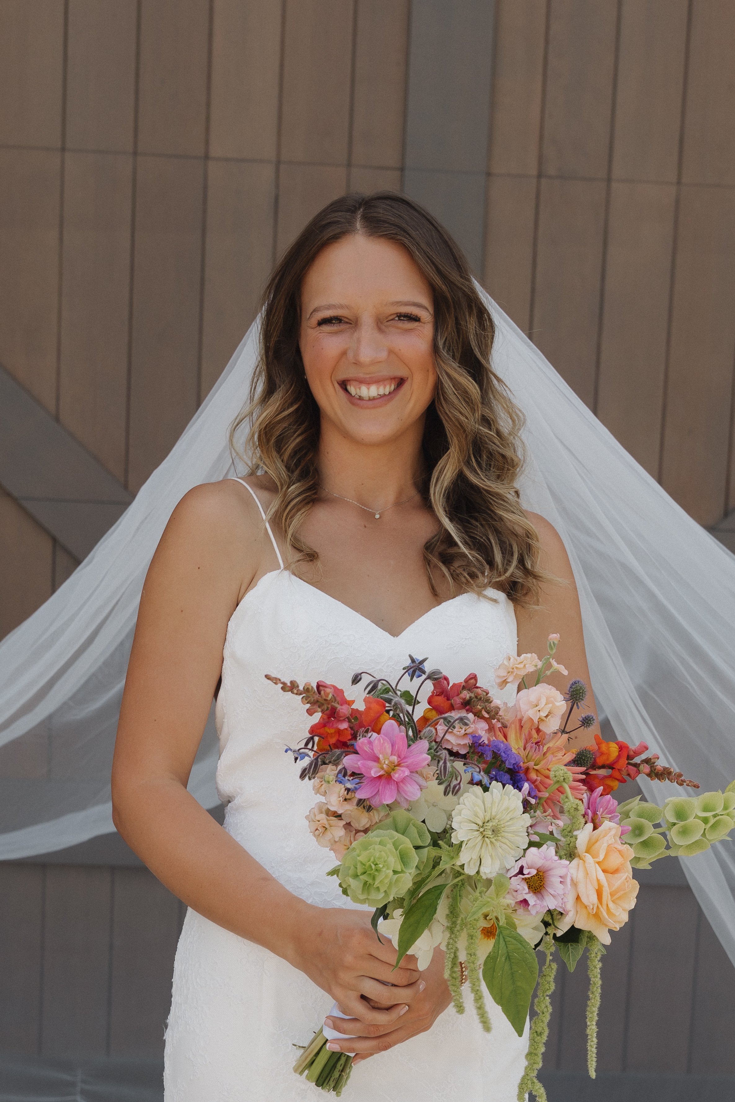 A smiling bride in a white wedding dress holding a colorful bouquet of flowers, standing in front of a modern building with a wooden exterior. She wears a veil and a delicate necklace.