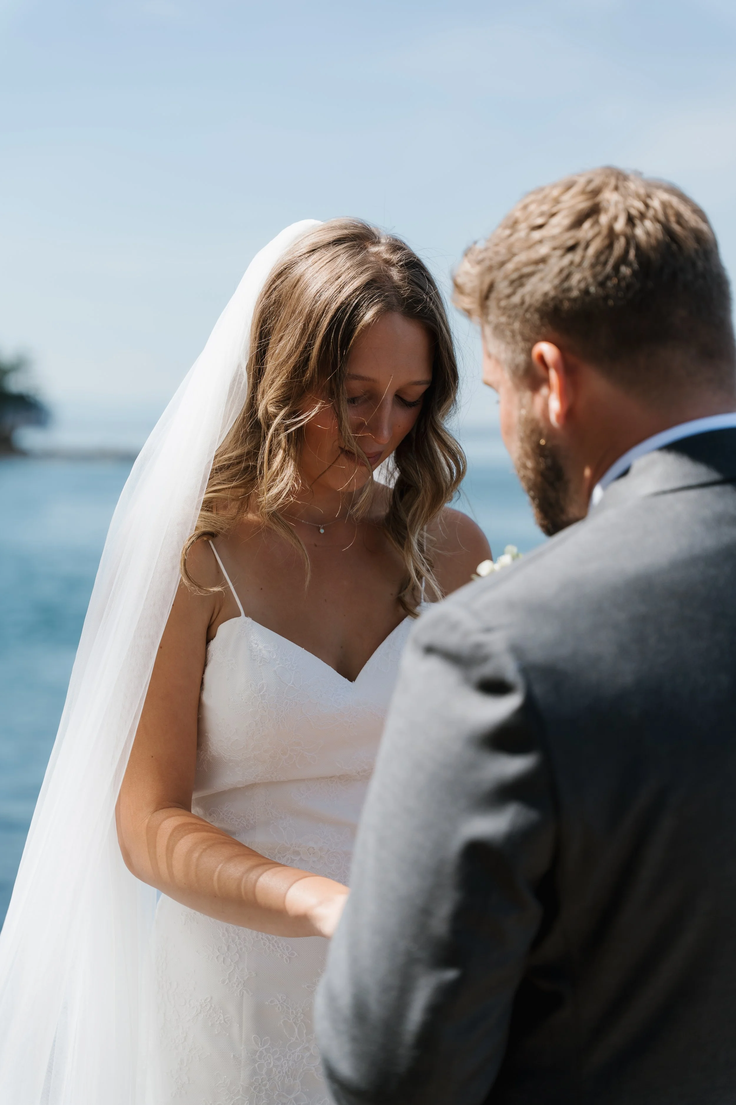 A bride and groom exchanging vows outdoors near a body of water, with the bride wearing a white wedding dress and veil, and the groom in a gray suit.