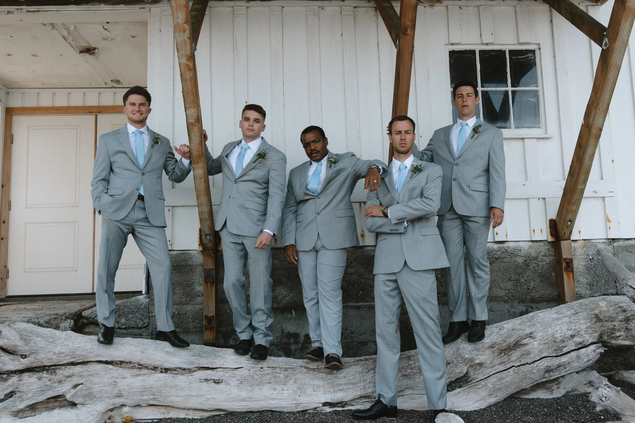Six men dressed in gray suits with light blue ties standing outdoors against a white wooden building with a window. They are posing confidently, some leaning or resting on wooden beams and a large piece of driftwood on the ground.