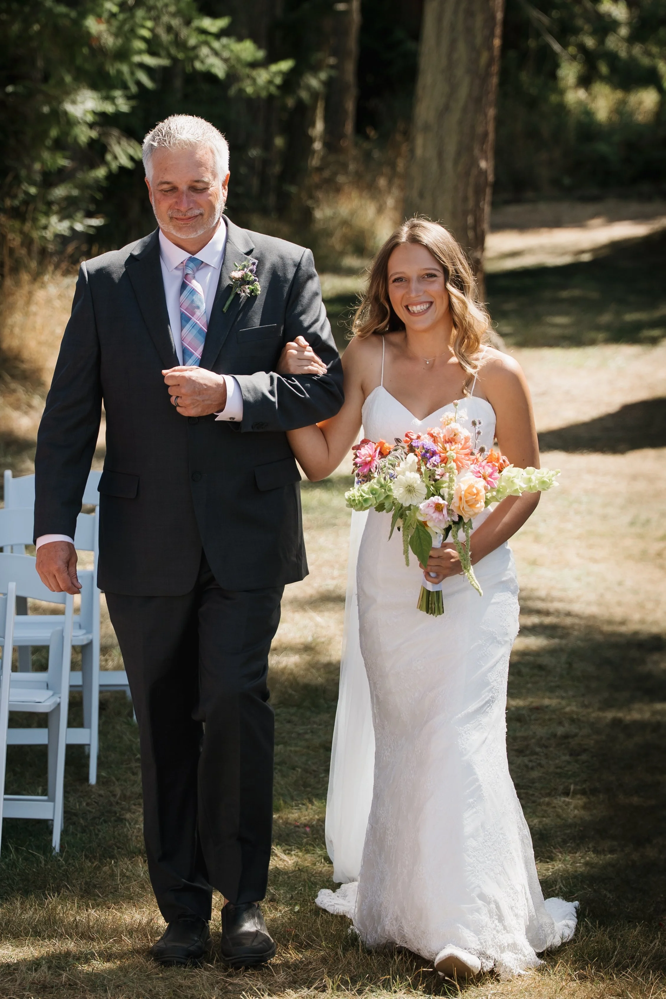 A bride walking outdoors arm-in-arm with a man, likely her father, on her wedding day. The bride is smiling and holding a bouquet of colorful flowers, wearing a white lace wedding dress. The man is dressed in a dark suit, white shirt, and colorful st