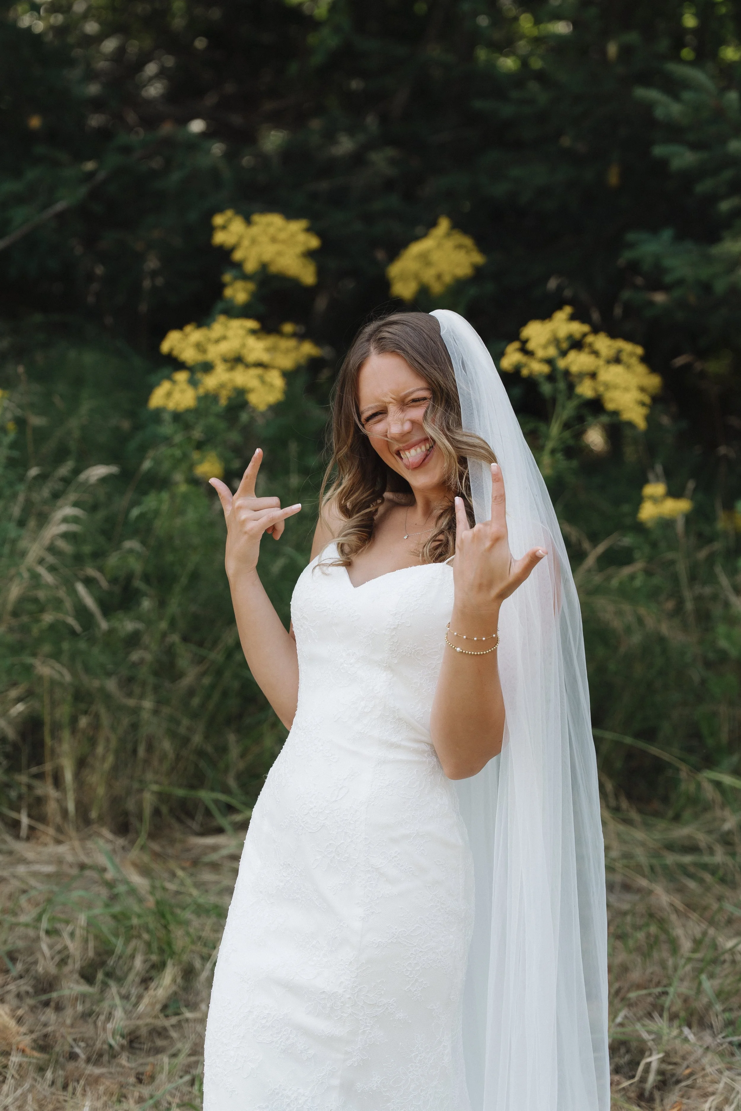 A woman in a white wedding dress and veil making rock and roll hand gestures and sticking out her tongue outdoors.