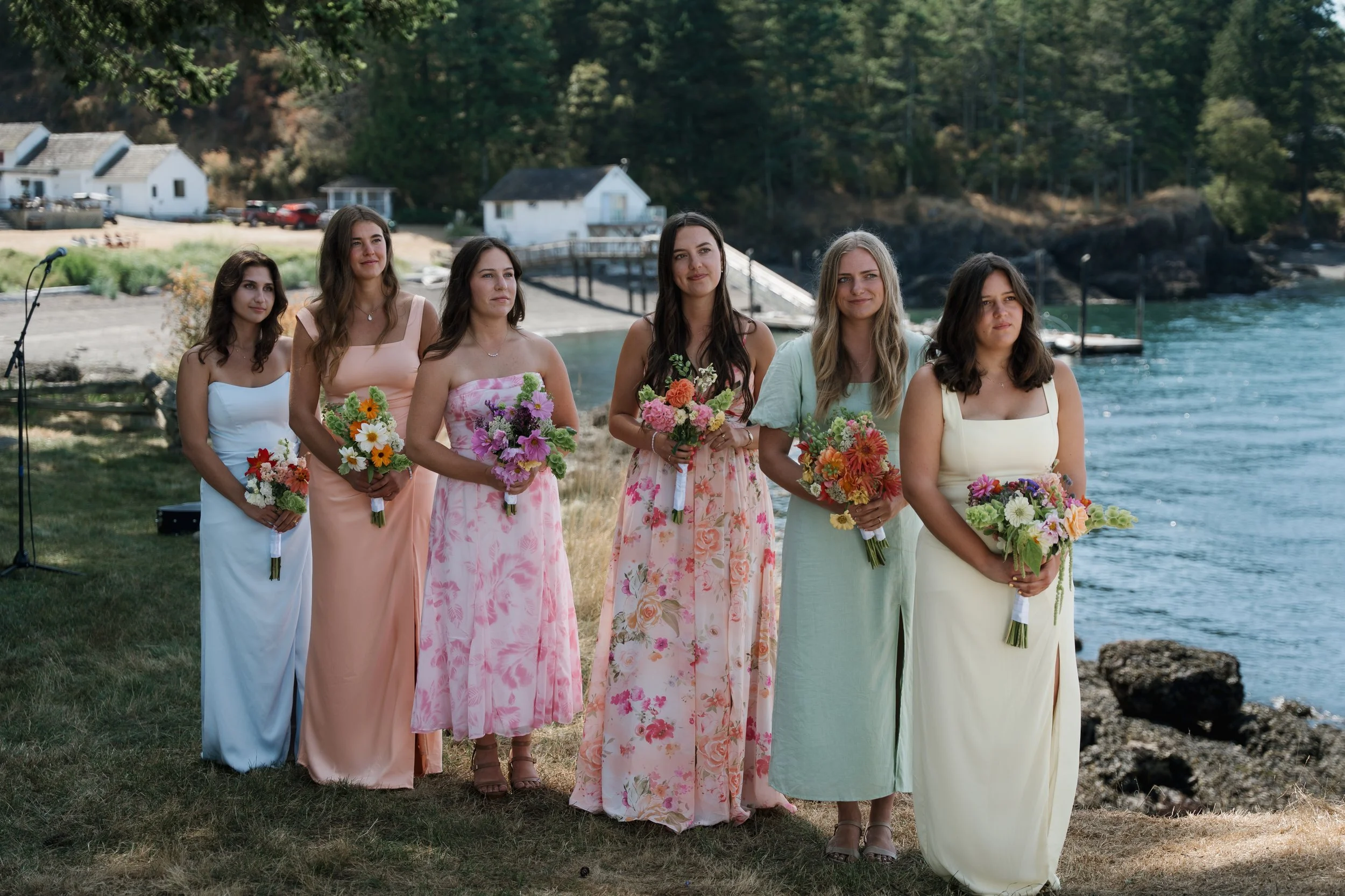 Six women dressed in pastel-colored dresses holding bouquets, standing outdoors near a body of water with houses and trees in the background.