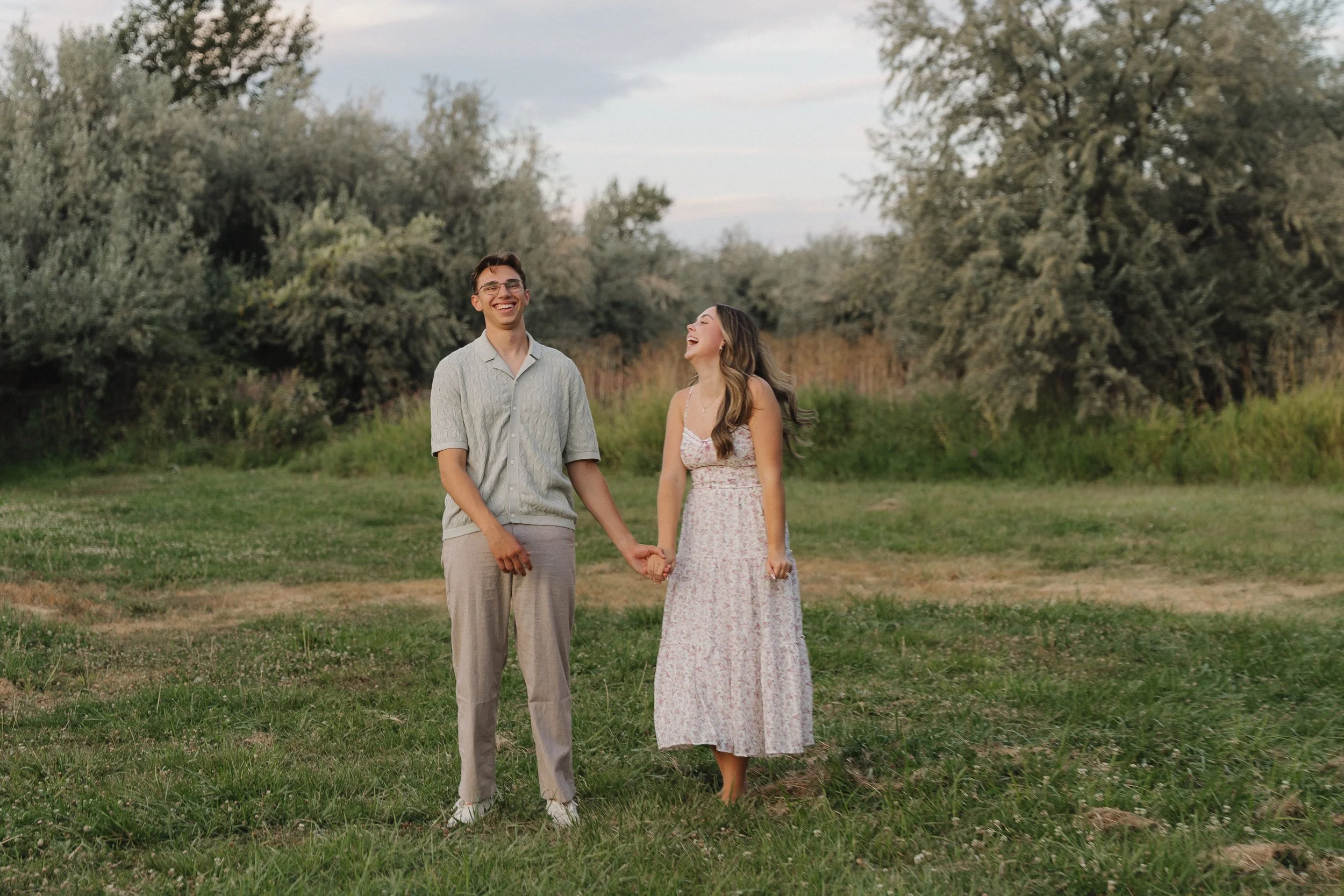A young couple holding hands and laughing in a grassy field with trees in the background