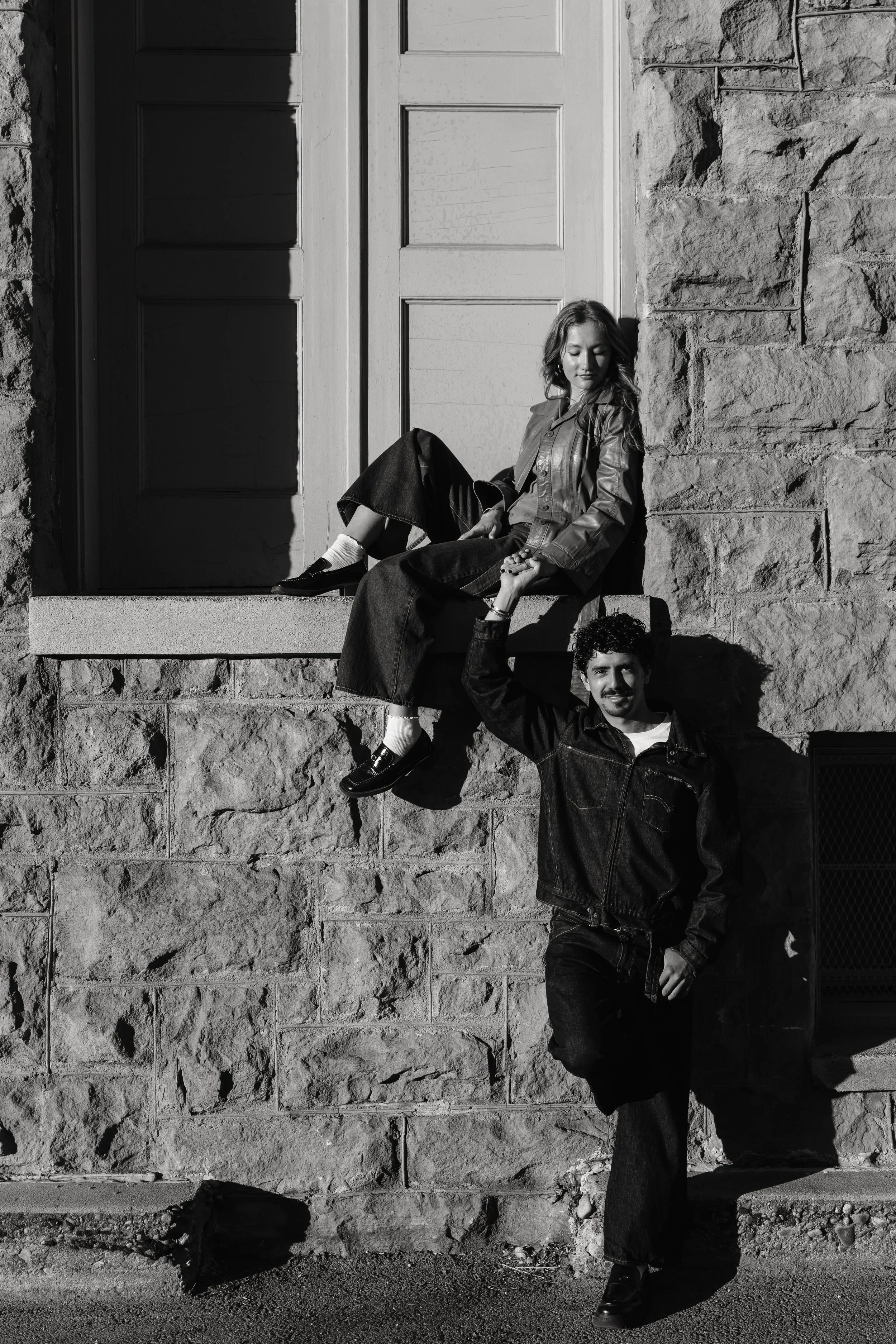 A black-and-white photo of a woman sitting on a ledge in front of a door and a man standing beside her, holding her hand, both smiling.