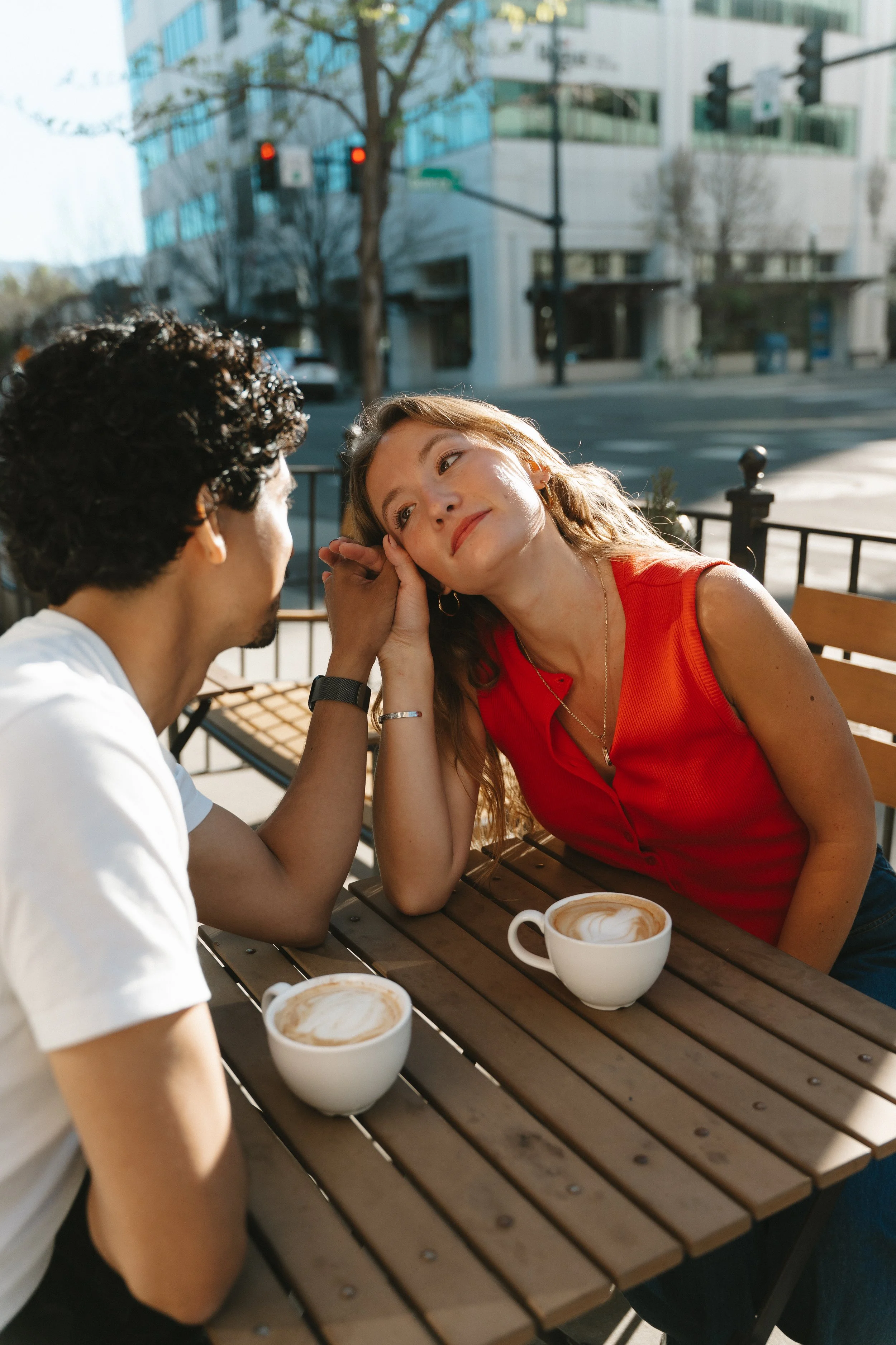 A man and woman sit at an outdoor cafe table with two cups of coffee, sharing an intimate moment as the woman leans in to rest her head on her hand, gazing at the man.