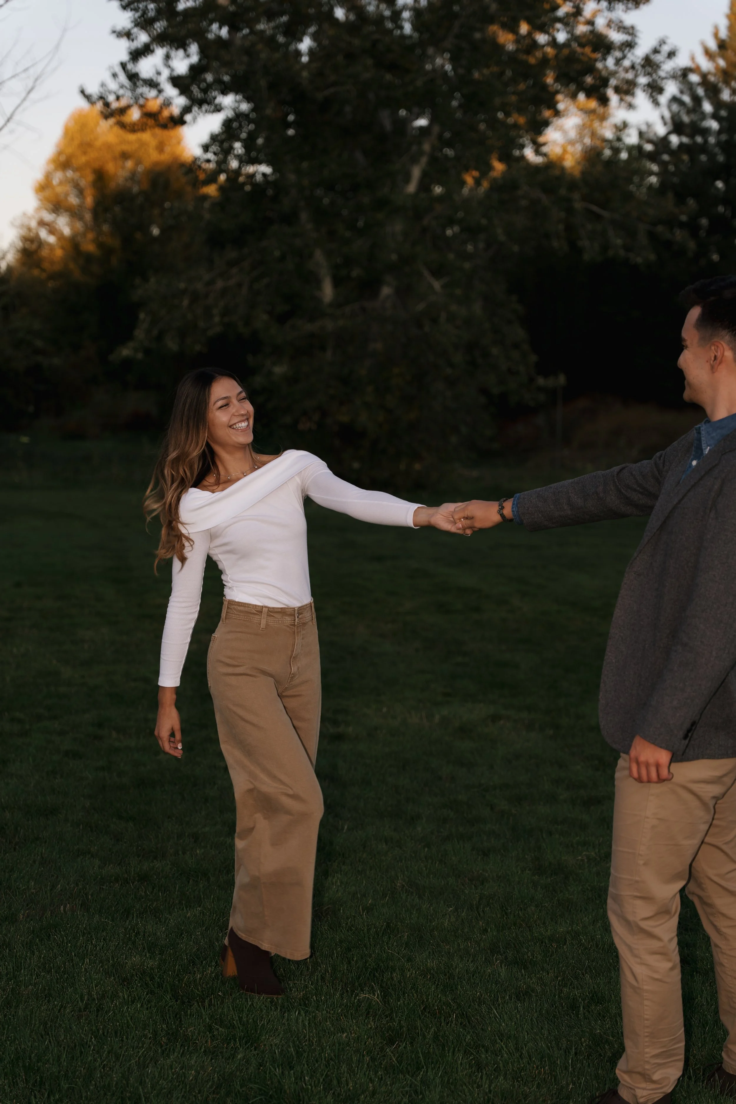 A woman and a man are smiling and holding hands outdoors on a grassy area during sunset, with trees in the background.