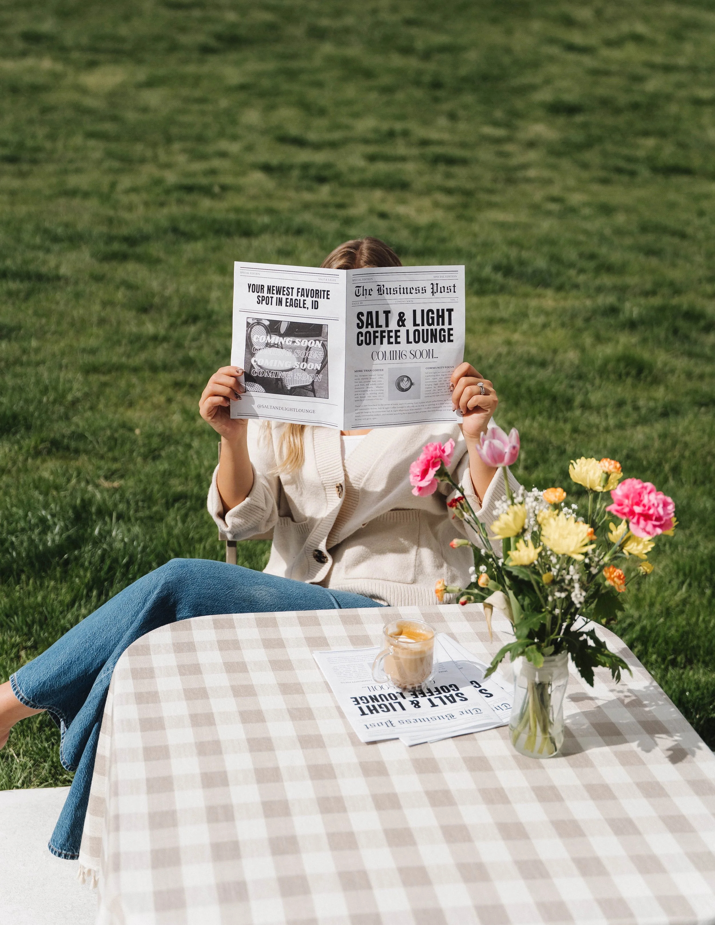 Woman sitting at a checkered table outdoors on grass, reading a newspaper. The table has a vase of pink and yellow flowers, a glass of iced coffee, and a newspaper. The woman is wearing a white cardigan and blue jeans.