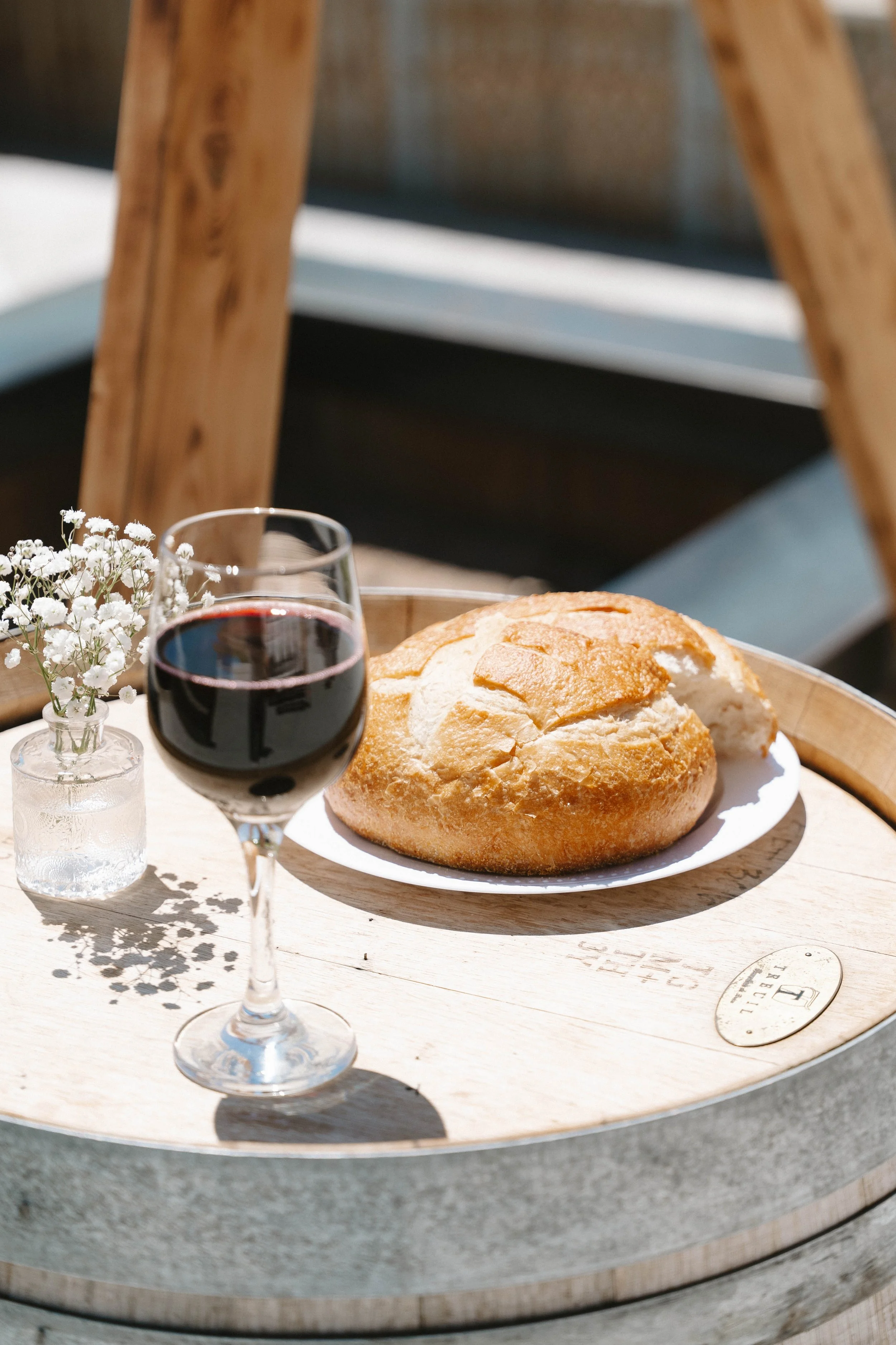 A glass of red wine, a loaf of bread on a white plate, and a small vase with white flowers on a wooden table outdoors.