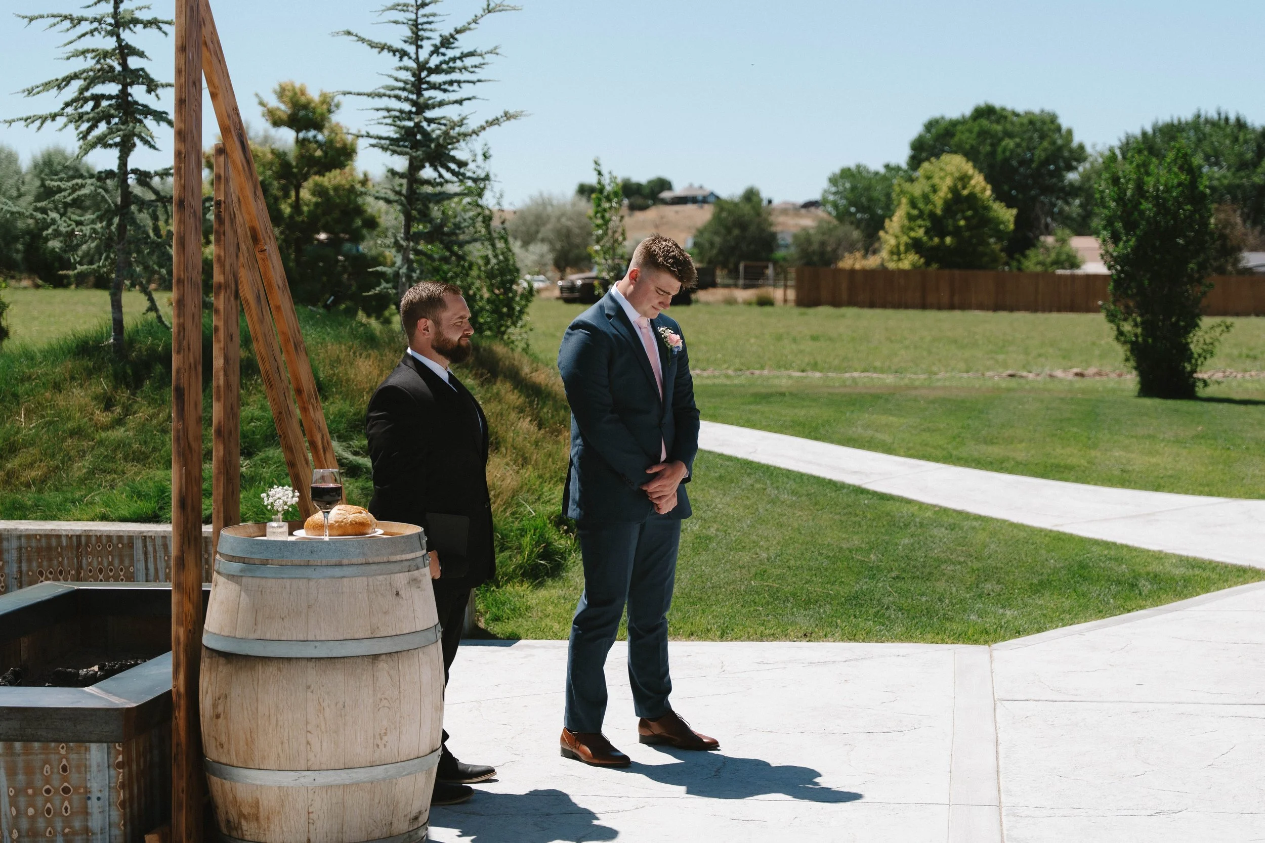 Two men in suits standing outdoors during a wedding ceremony, with one praying and the other bowing head, near a decorated table with bread, wine, and flowers