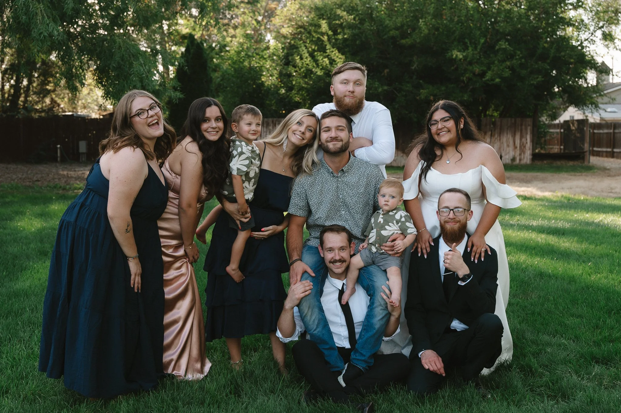 Group of people, including adults and children, posing outdoors on a grassy lawn with trees and a fence in the background, smiling and dressed in semi-formal attire.