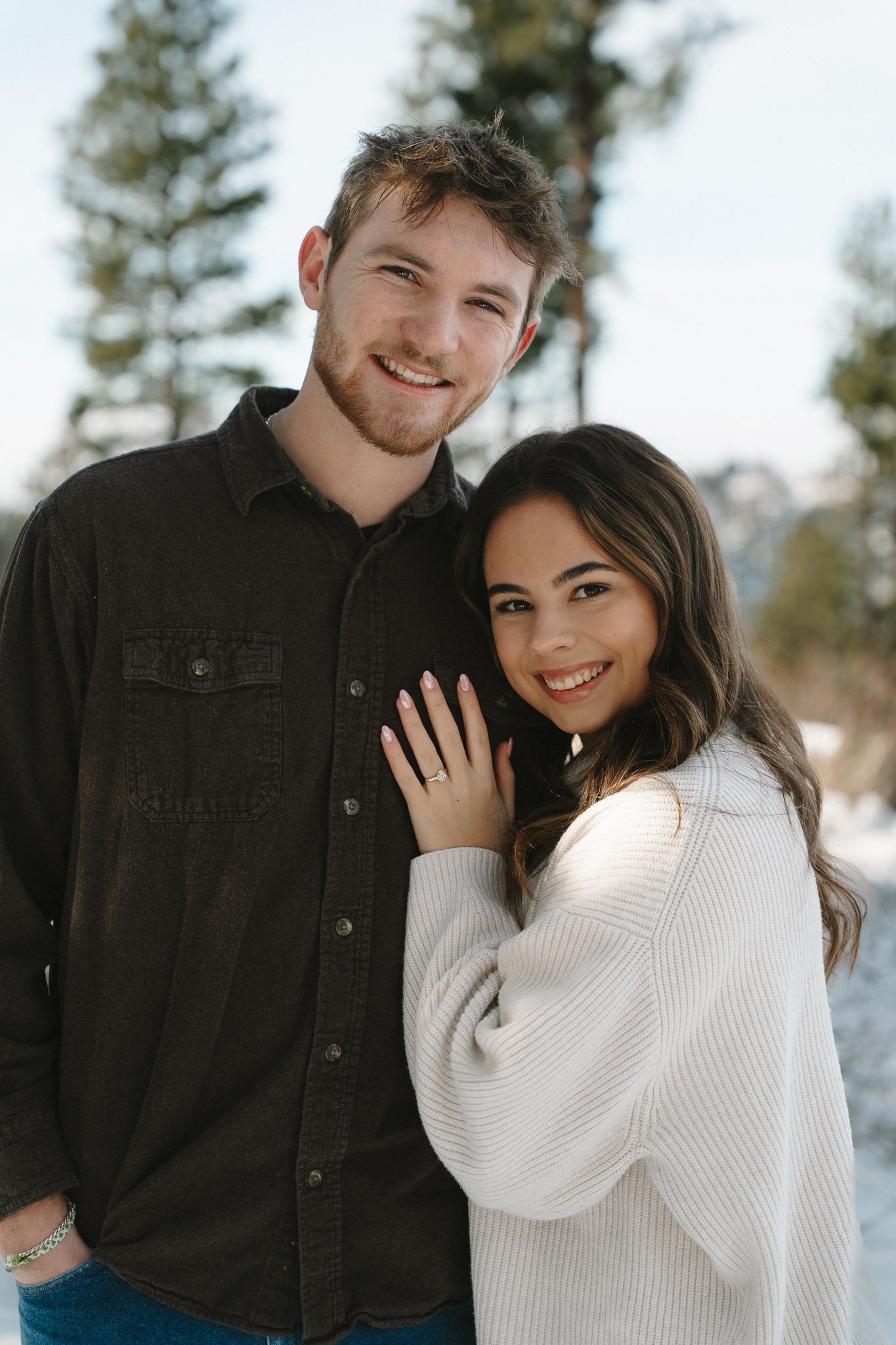 A young woman with long dark hair and a white sweater is smiling with her hand on the chest of a smiling young man with short brown hair and a black denim jacket. They are outdoors with trees in the background.