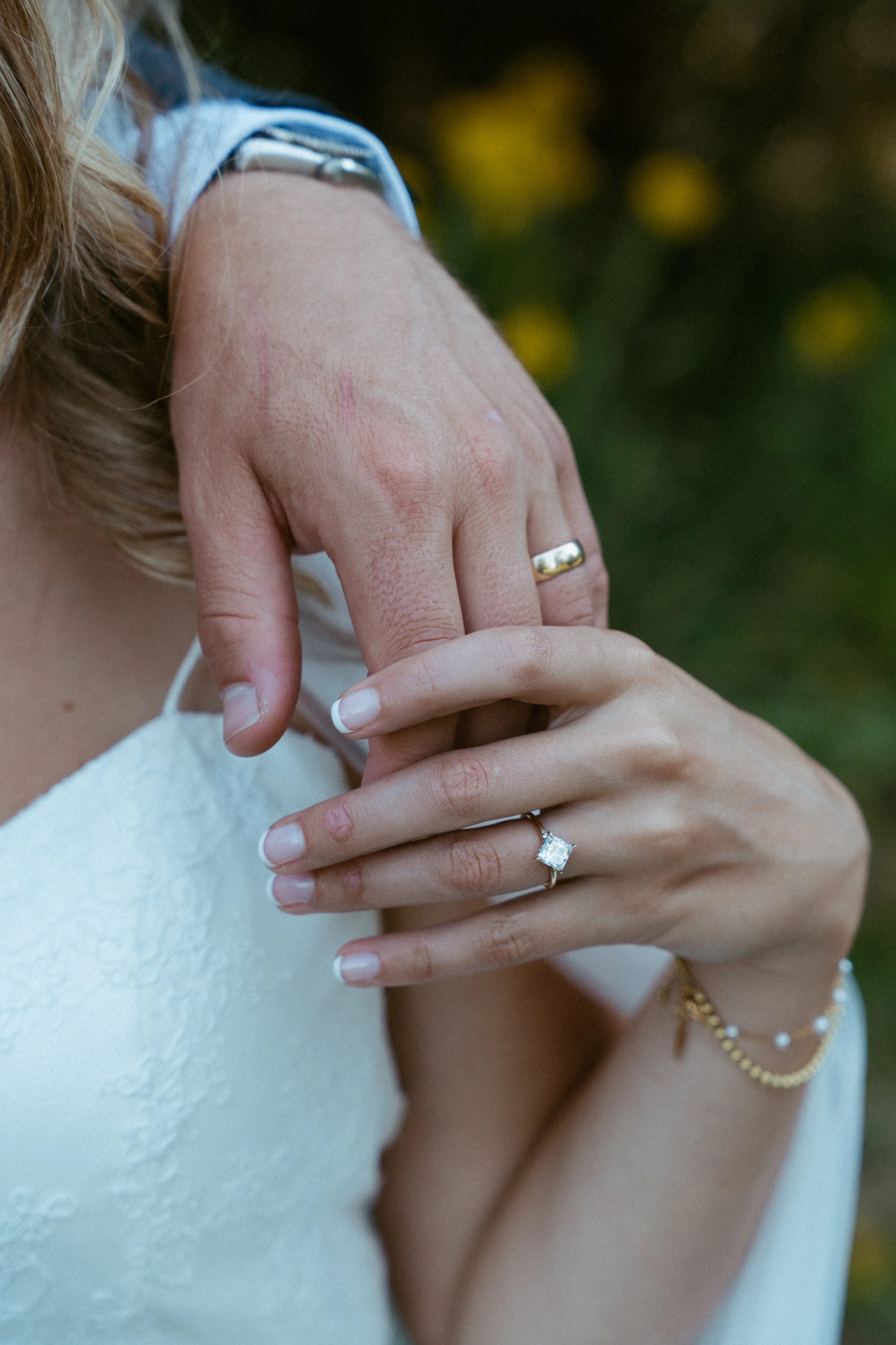 Close-up of a bride and groom's hands showing wedding rings, with the groom's hand on top of the bride's hand, against a blurred outdoor background.