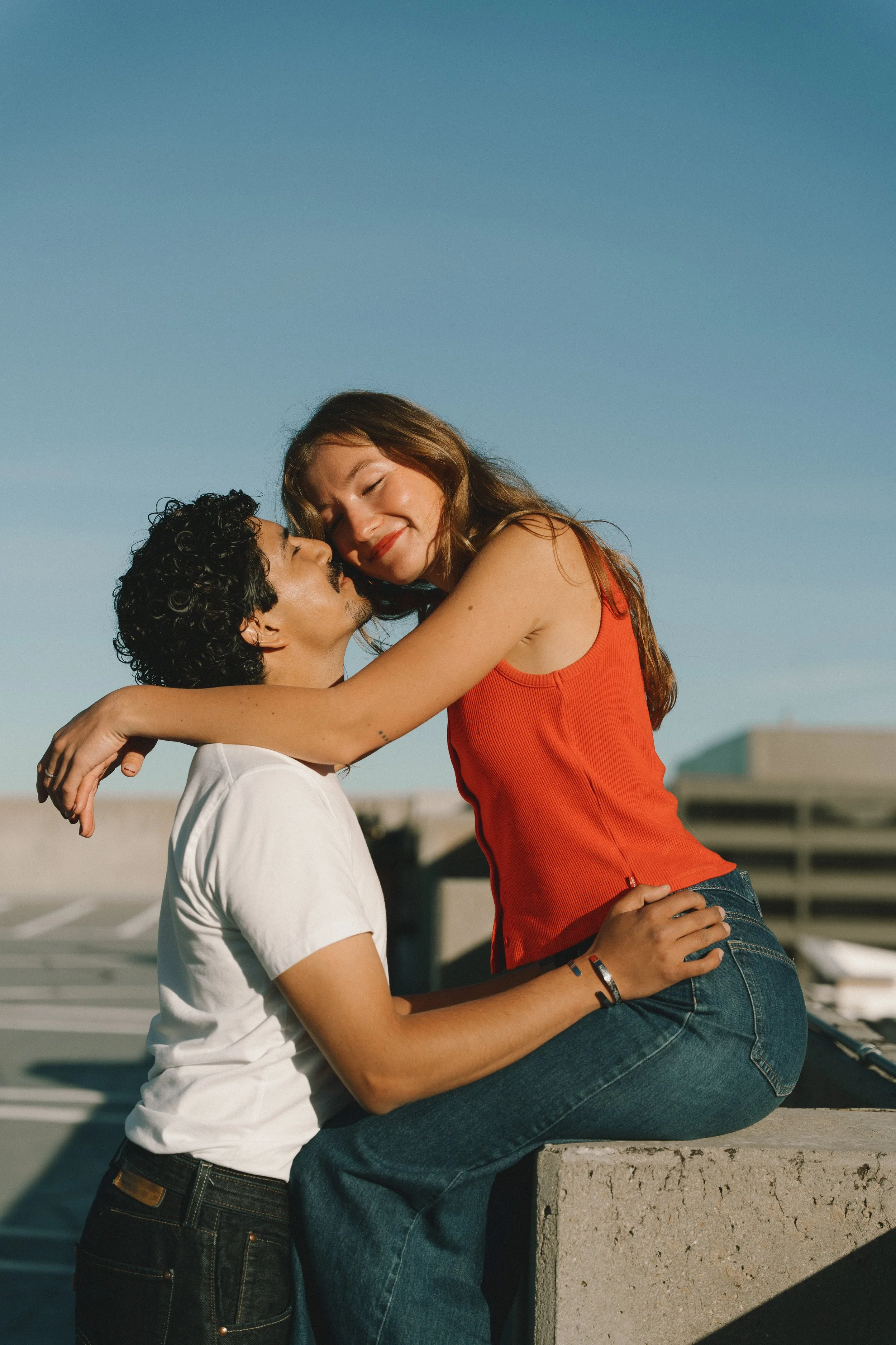 A young couple happily embracing on a rooftop, with a clear blue sky in the background.
