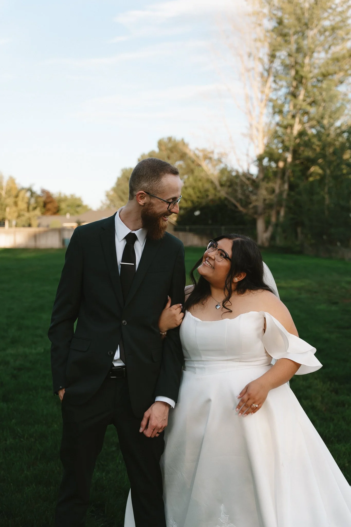 A newlywed couple standing arm in arm outdoors on a grassy field, smiling at each other. The groom wears a black suit with a white shirt and black tie, and the bride wears a white off-shoulder wedding dress with ruffled sleeves and glasses. Trees and