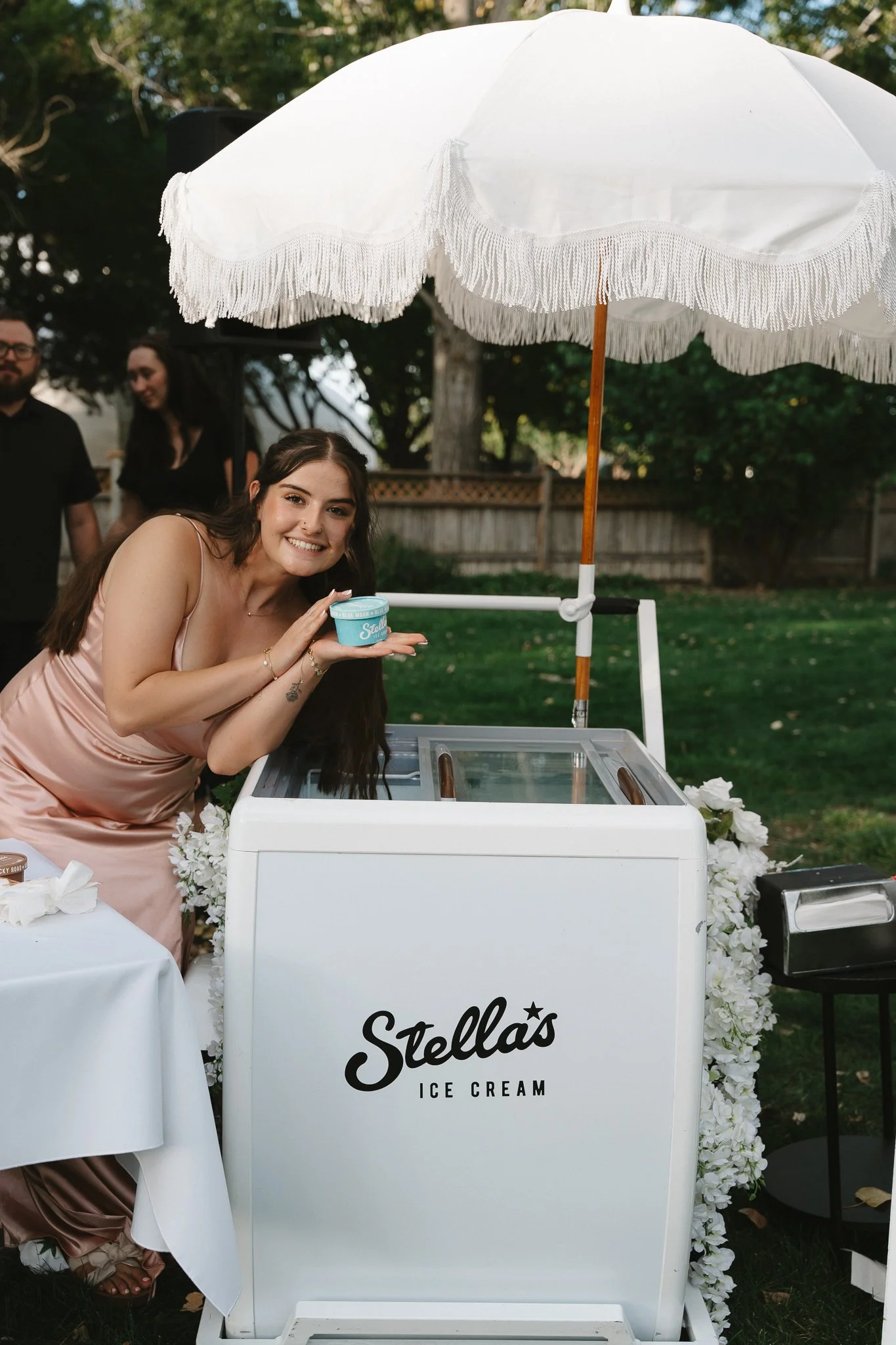 A young woman in a silky pink dress is smiling and holding a small tub of Stella's ice cream while leaning on an ice cream cart under a white umbrella, with trees and a wooden fence in the background.