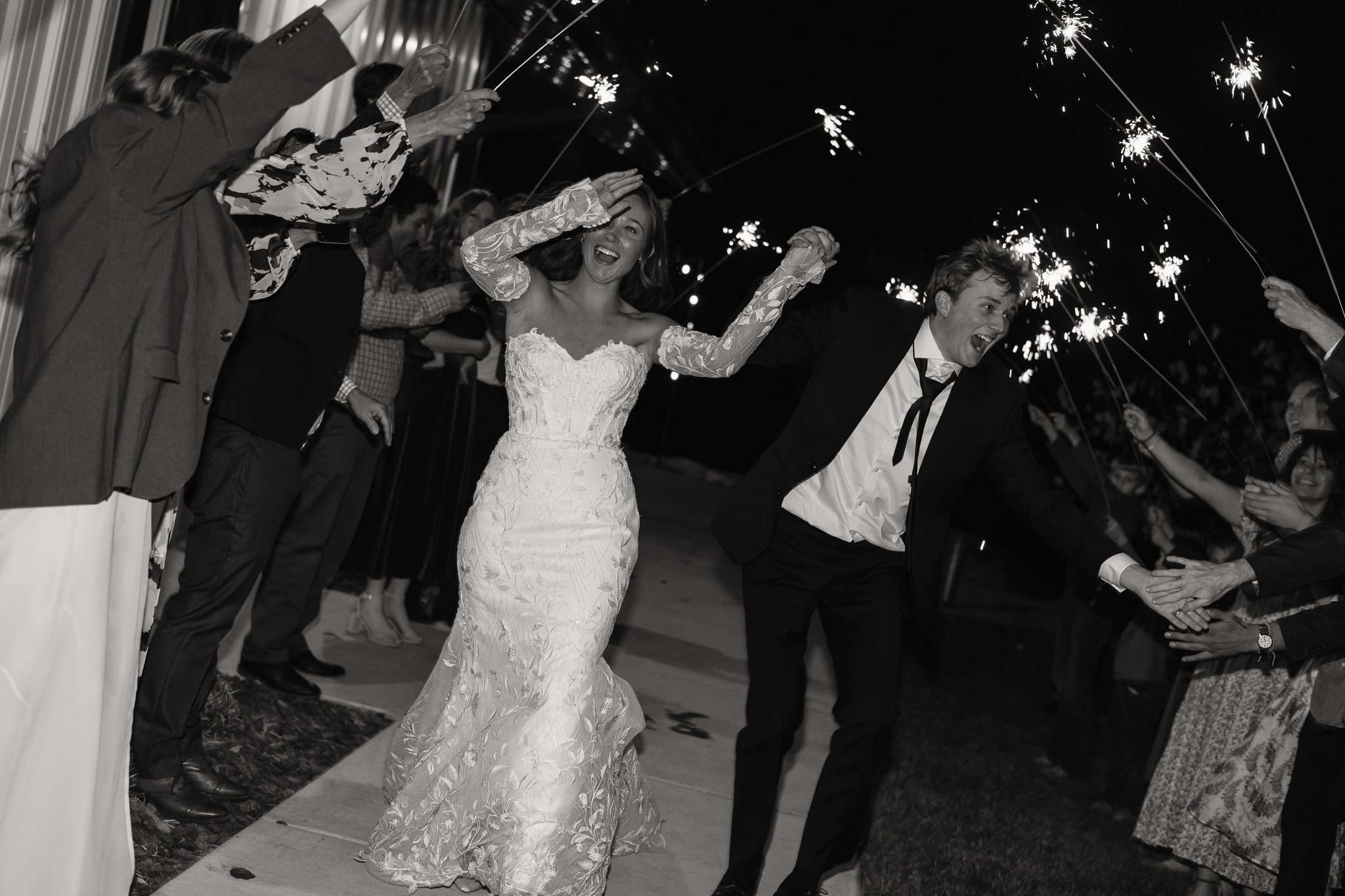 A newlywed couple celebrates with sparklers surrounded by friends and family at night.