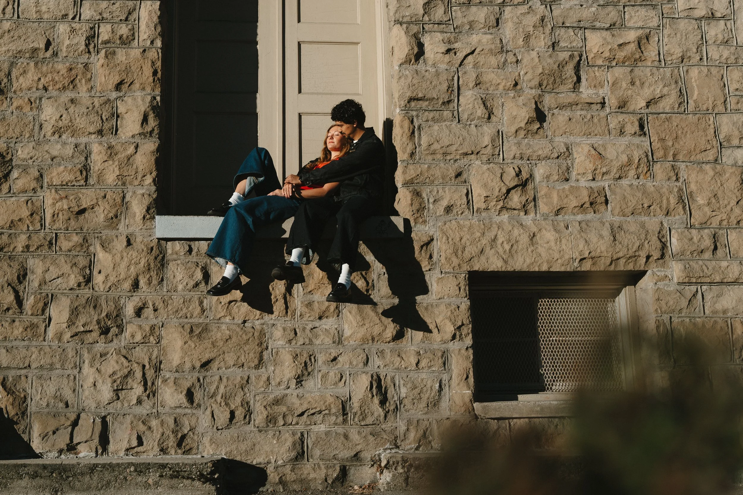 Two young adults, a man and a woman, sitting on a window ledge of a brick building, sharing a quiet moment in the sunlight, with shadows cast by the sunlight.