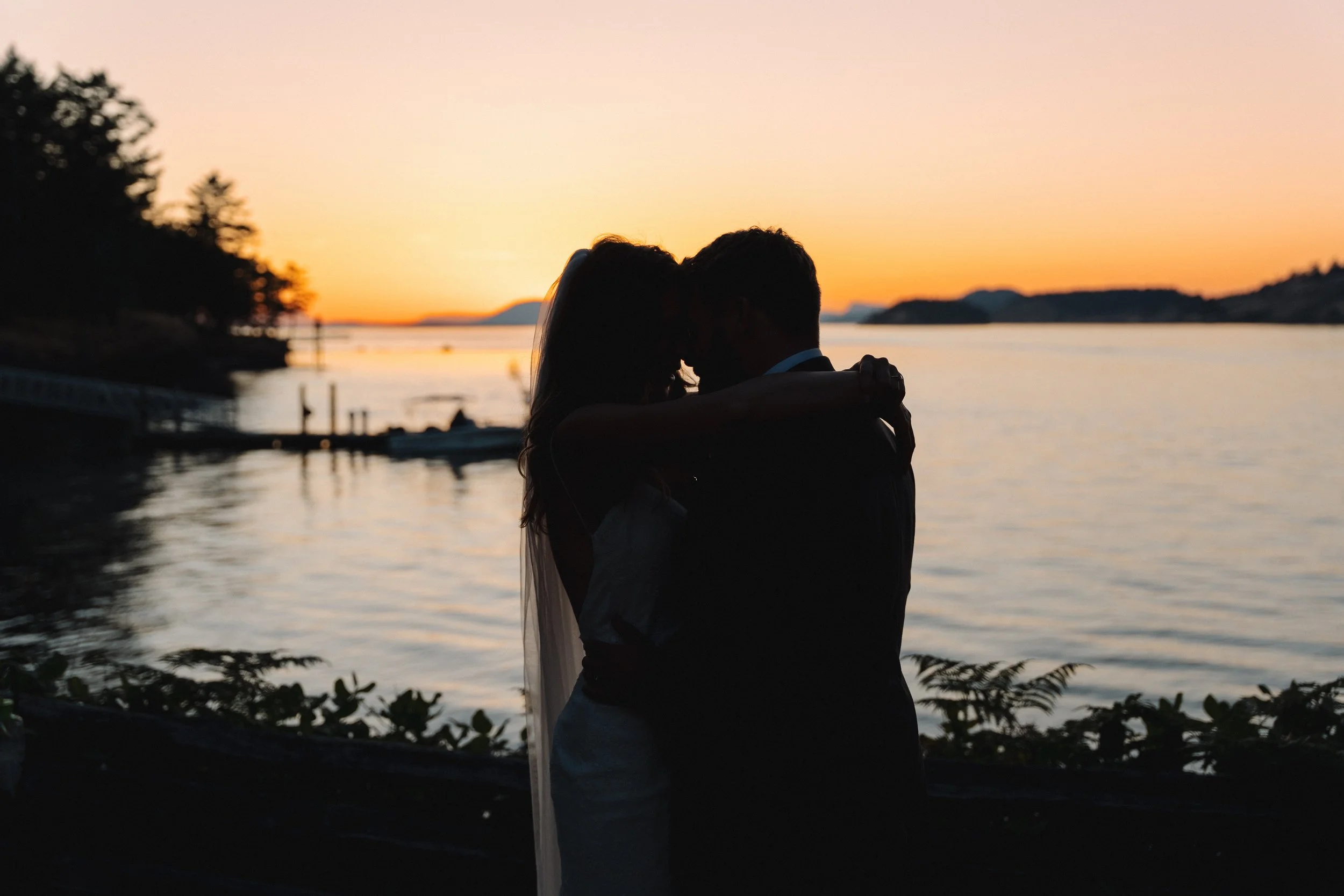 A couple embracing by the water at sunset, with silhouettes of trees and boat in the background.