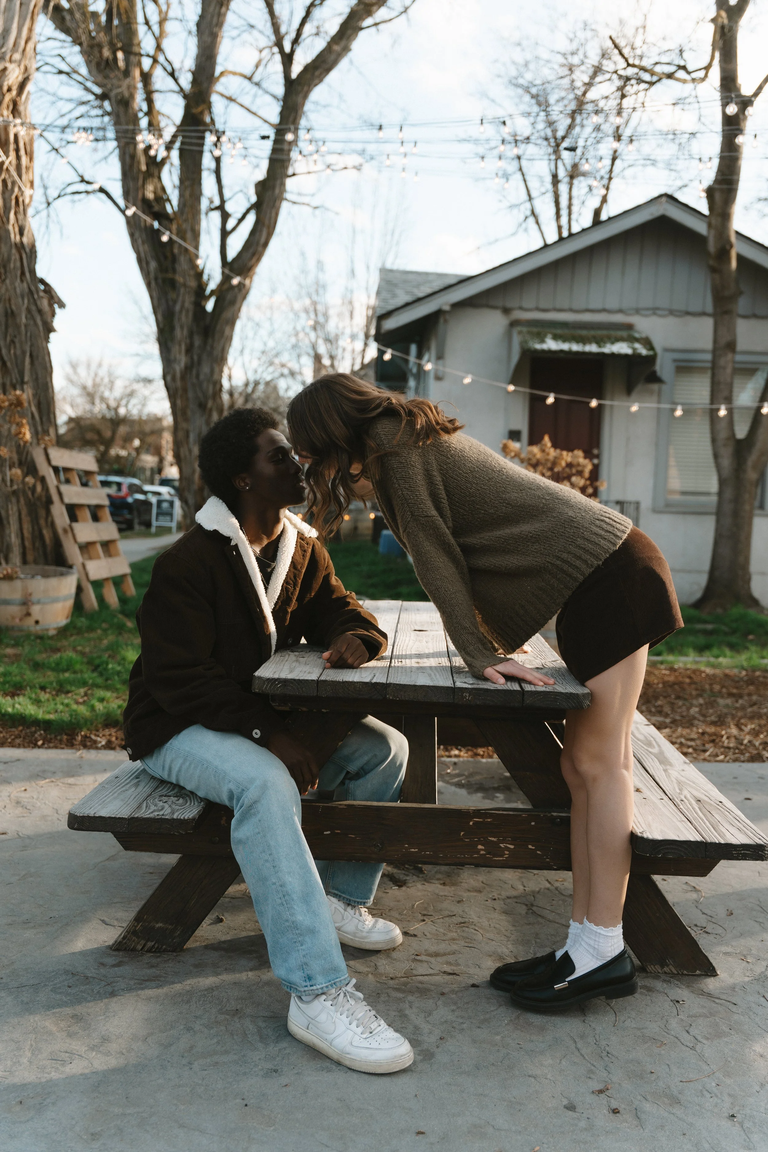 Two women are at a park on a picnic table, leaning towards each other for a kiss amid outdoor string lights, trees, and a house in the background.