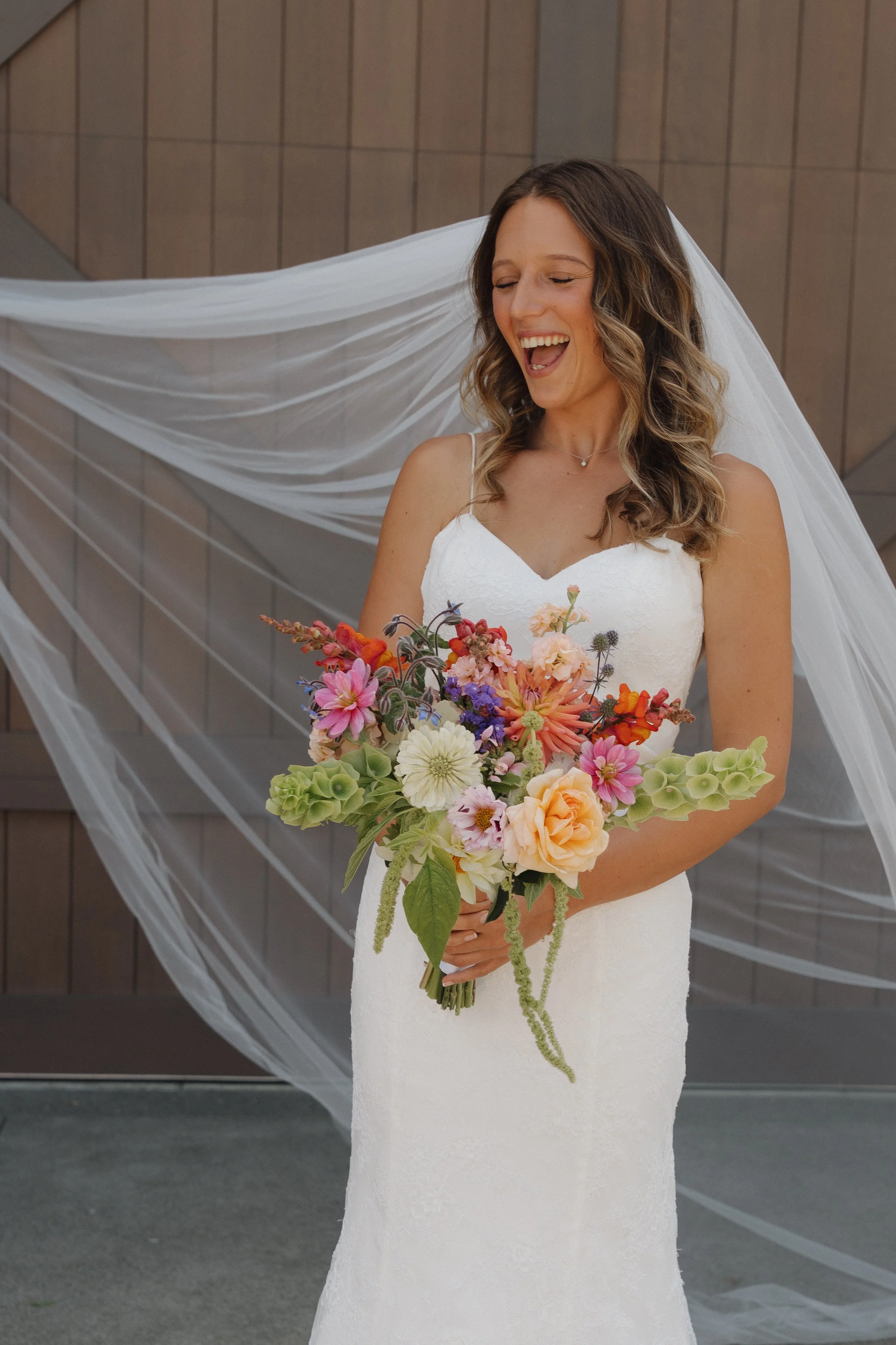 A bride with brown wavy hair in a white lace wedding dress, holding a vibrant bouquet of pink, orange, purple, and white flowers, smiling with her eyes closed.