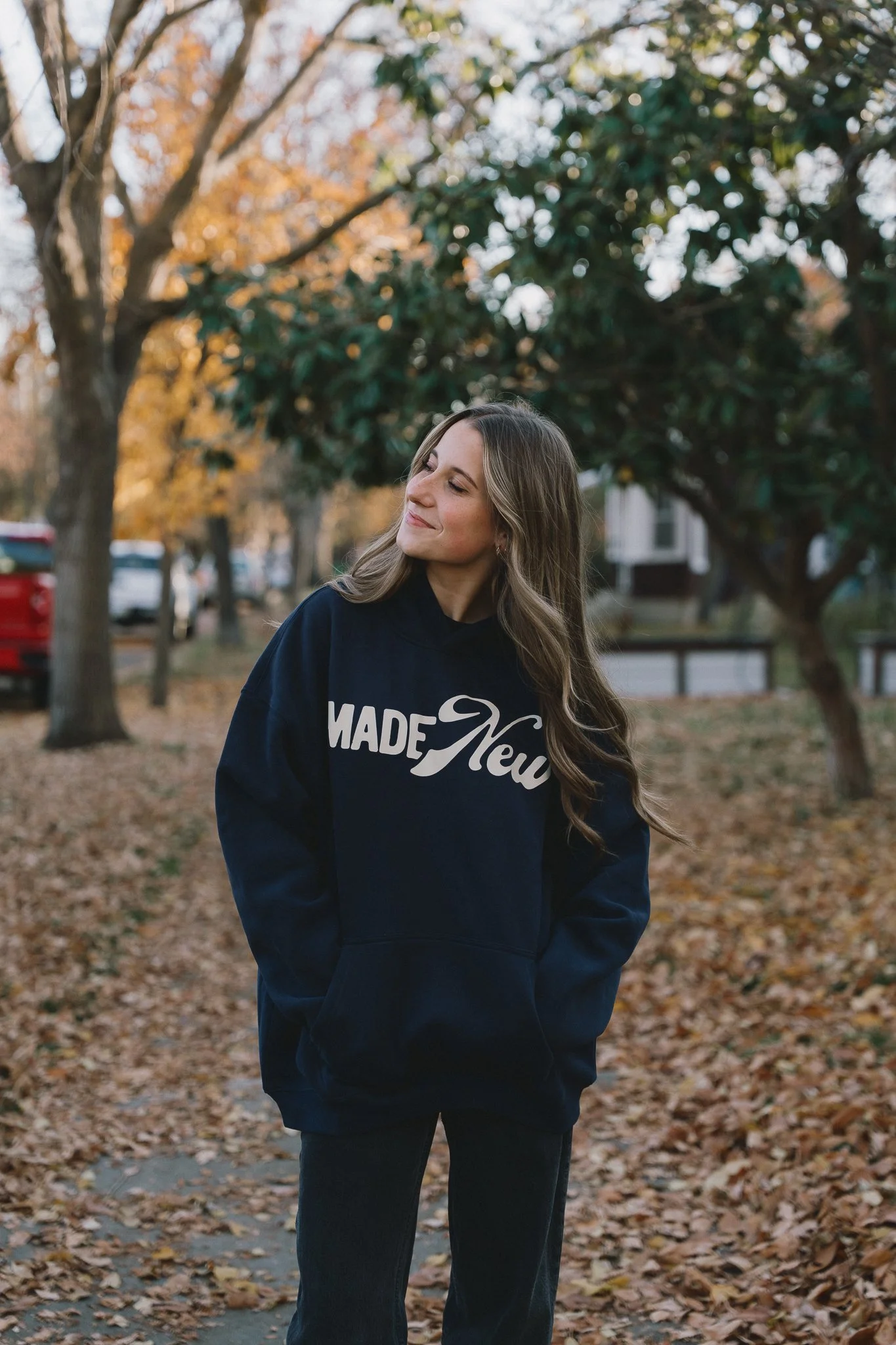 A woman in a navy sweatshirt with 'MADE New' written on it stands outdoors on a fall day, surrounded by trees with orange and yellow leaves.