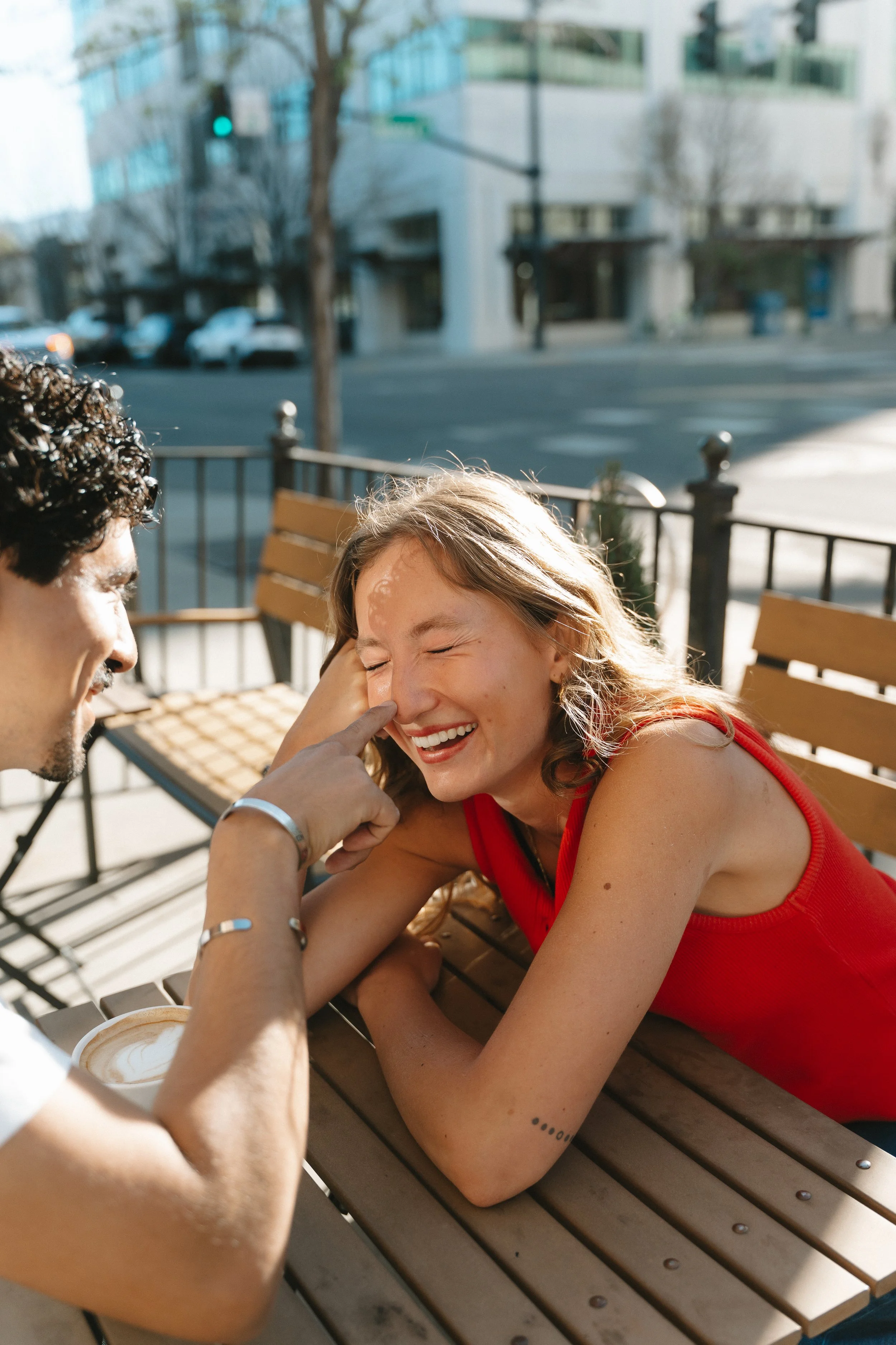 A young woman with blonde hair in a red sleeveless top sitting at an outdoor wooden table, laughing as a young man with dark hair playfully touches her nose.