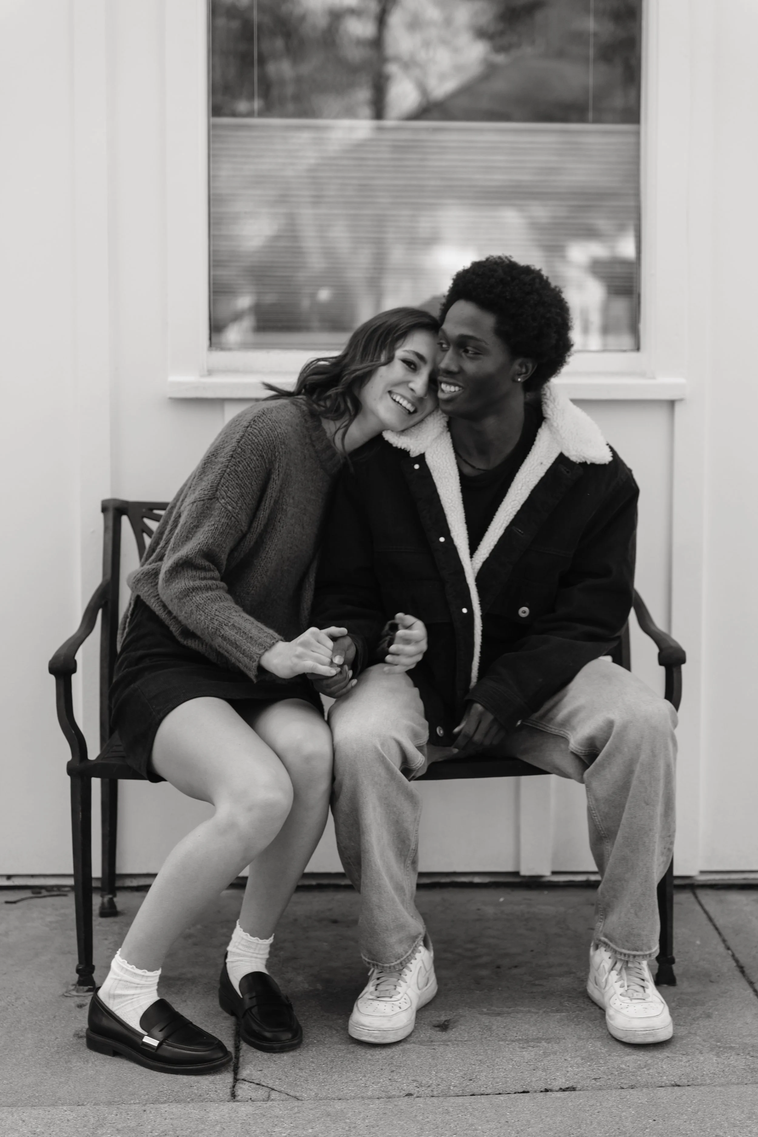 A young woman and man sitting closely on a porch bench, smiling and leaning their heads together. She has shoulder-length hair and is wearing a sweater, skirt, white socks, and loafers. He has short curly hair and is dressed in a jacket, hoodie, and 