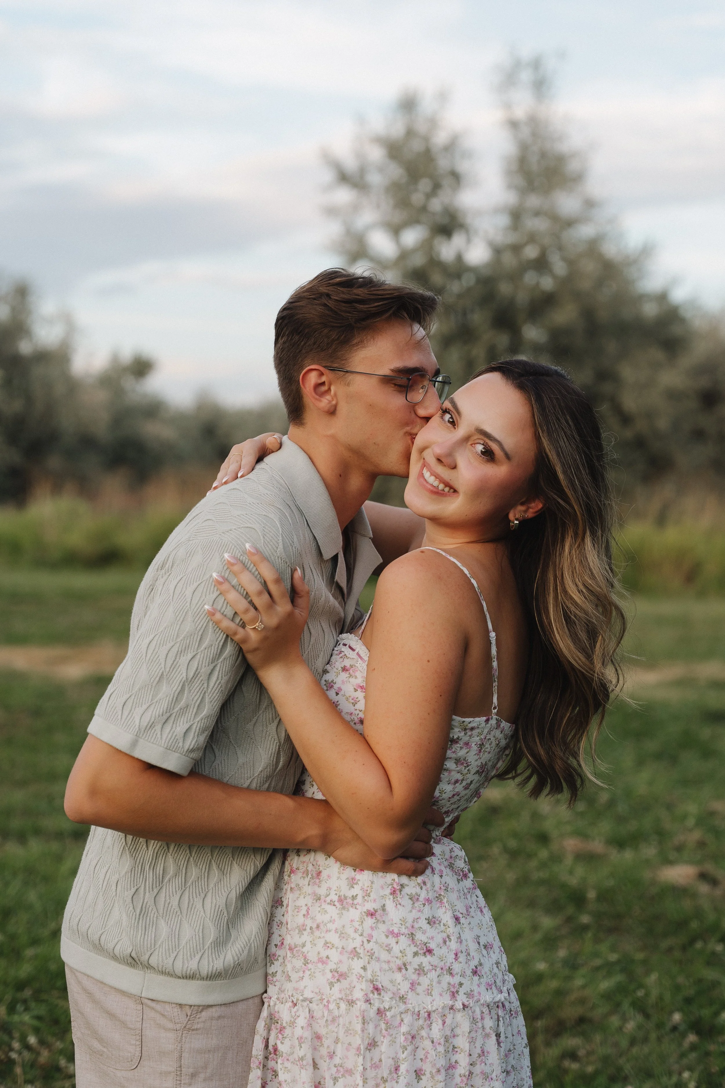 A young couple hugs outdoors with the man kissing the woman's cheek, both smiling, during the daytime in a natural setting.