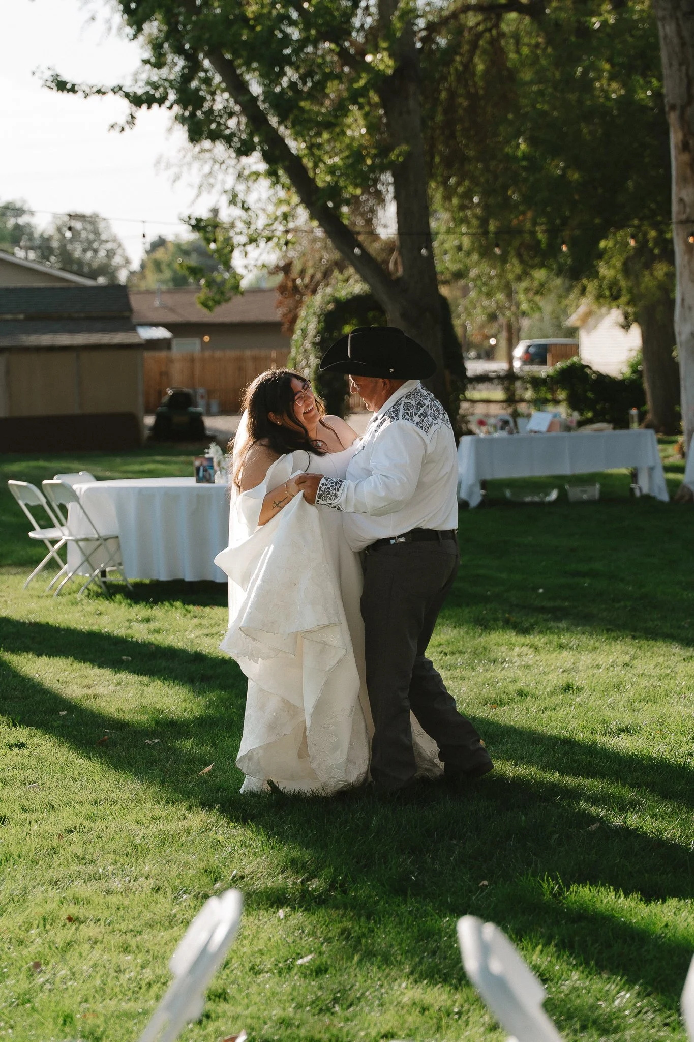A bride and a man with a cowboy hat dancing outdoors on a grassy area during daytime at a wedding celebration.