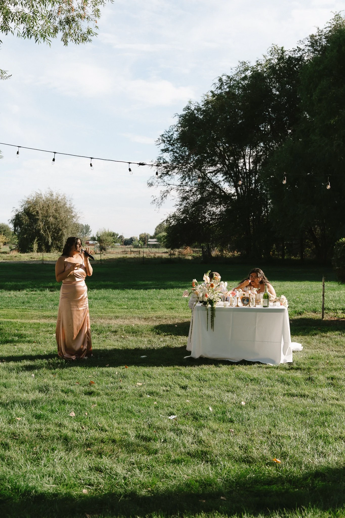 Two women at an outdoor event, one standing with a microphone and the other sitting at a decorated table, on a grassy field with trees and string lights overhead.