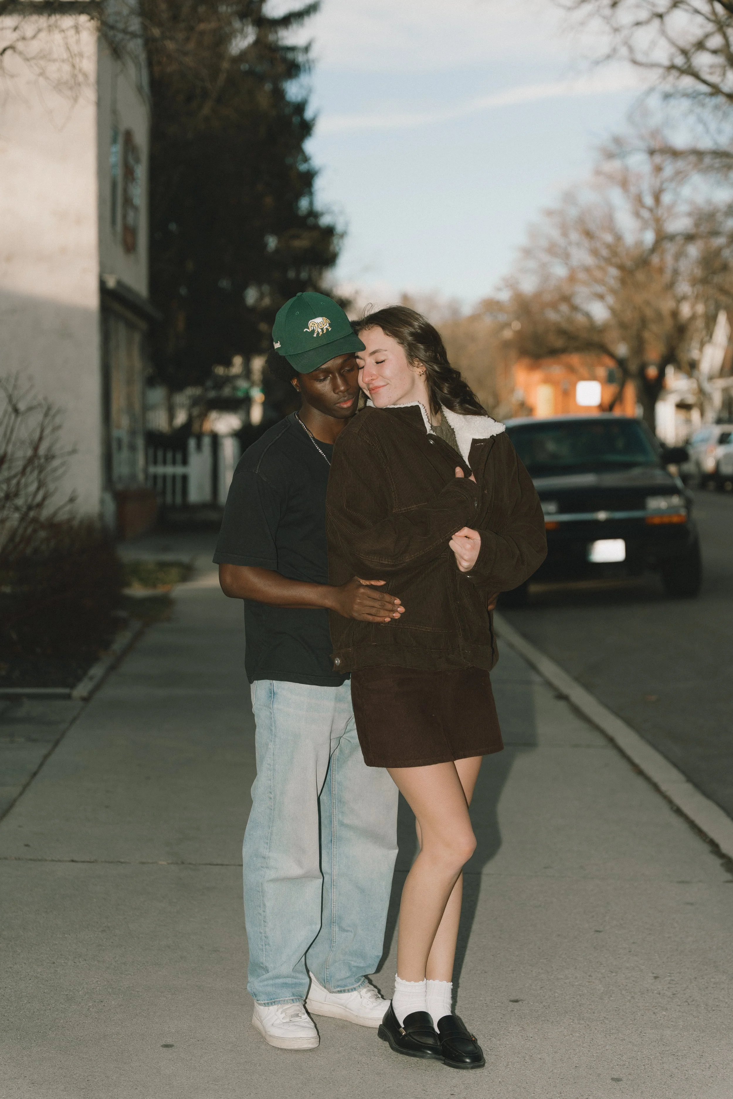 A young couple hugging on a sidewalk during the daytime, with parked cars and trees in the background.