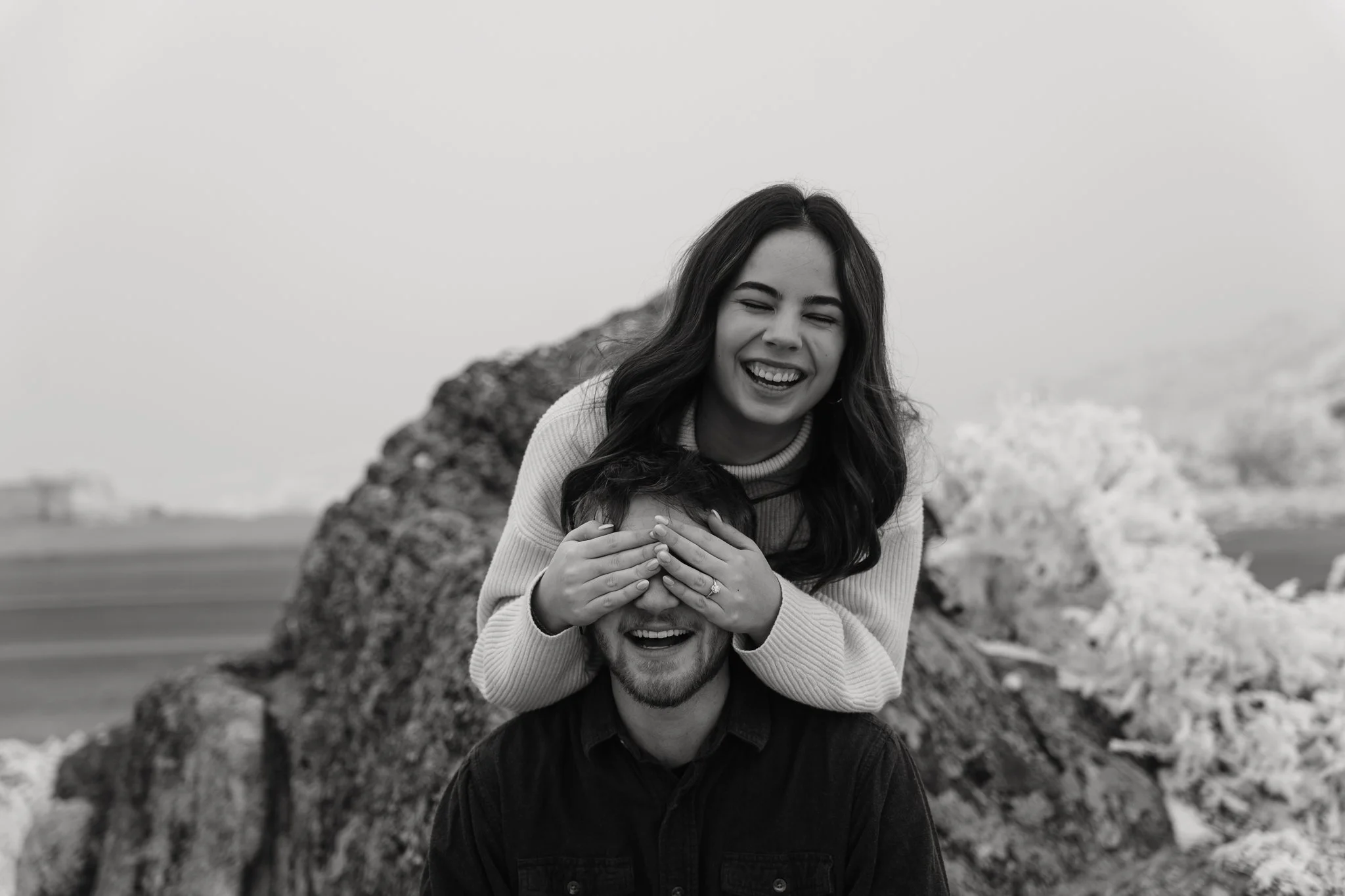 A woman playfully covers a man's eyes with her hands as they both smile, outdoors near rocks and water.