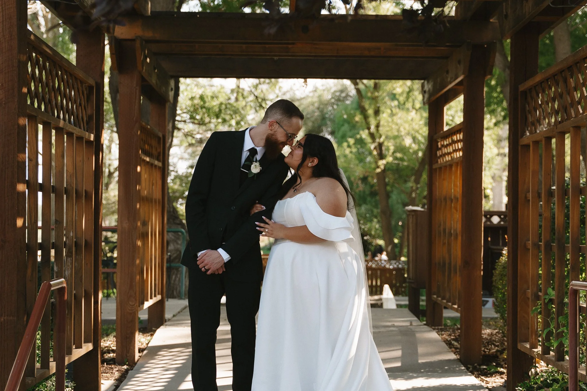 A bride and groom sharing a moment together under a wooden archway in an outdoor setting, with trees and sunlight in the background.