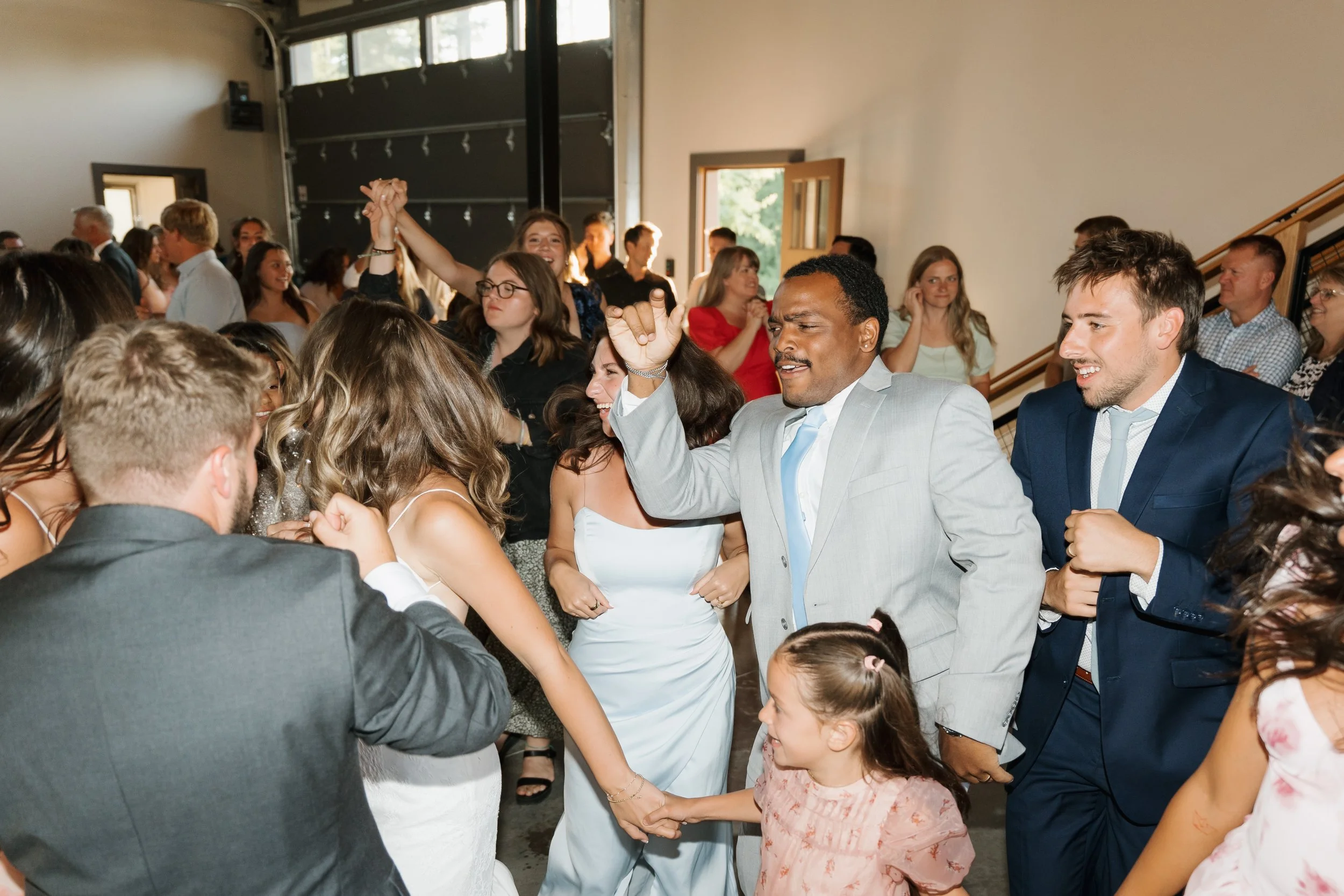 People dancing and celebrating at a wedding reception, with a woman in a white dress and a man in a light gray suit in the center.