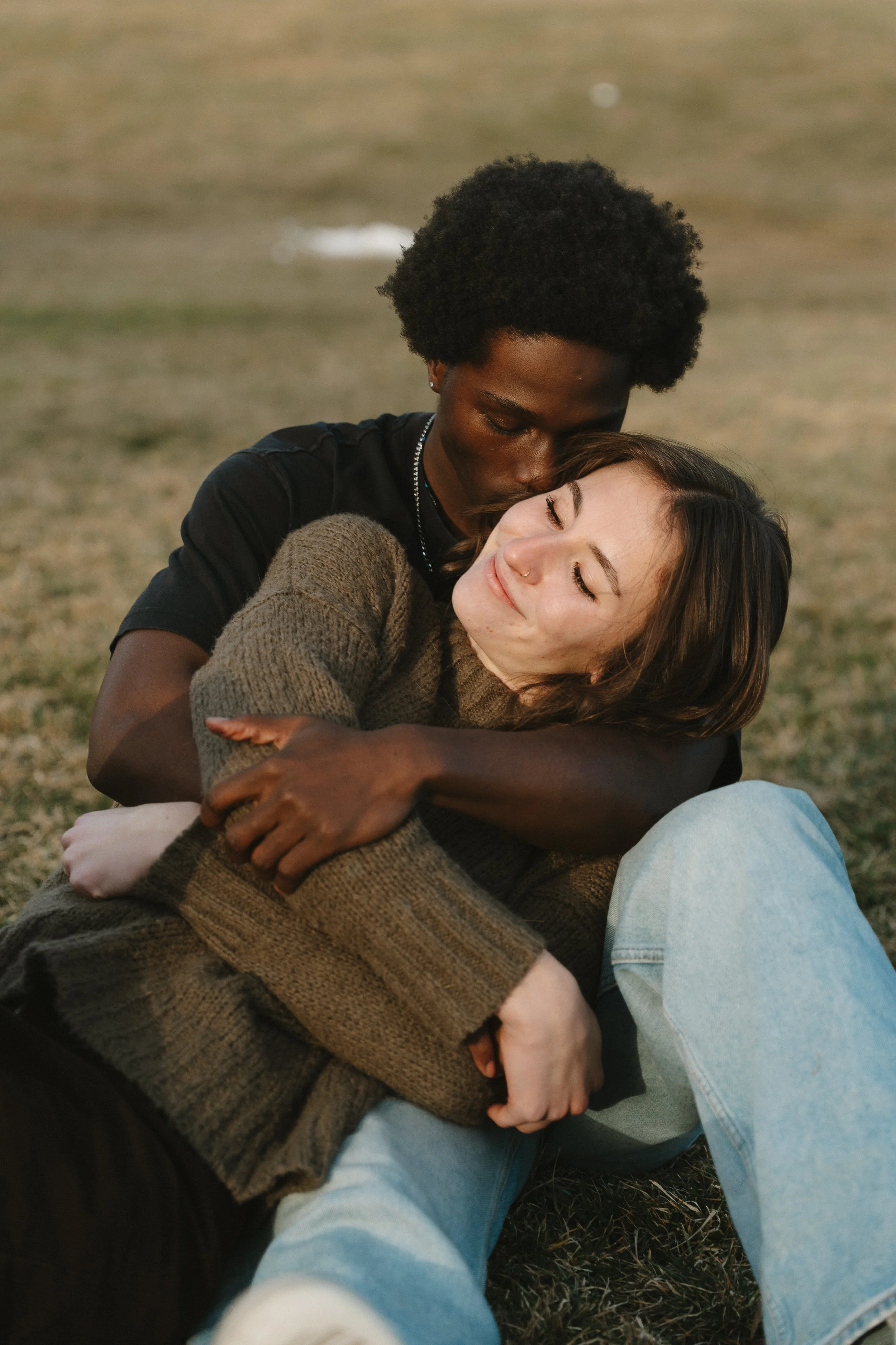 Two friends, one with dark skin and curly hair and the other with light skin and straight brown hair, sharing a hug outdoors on a grassy field.