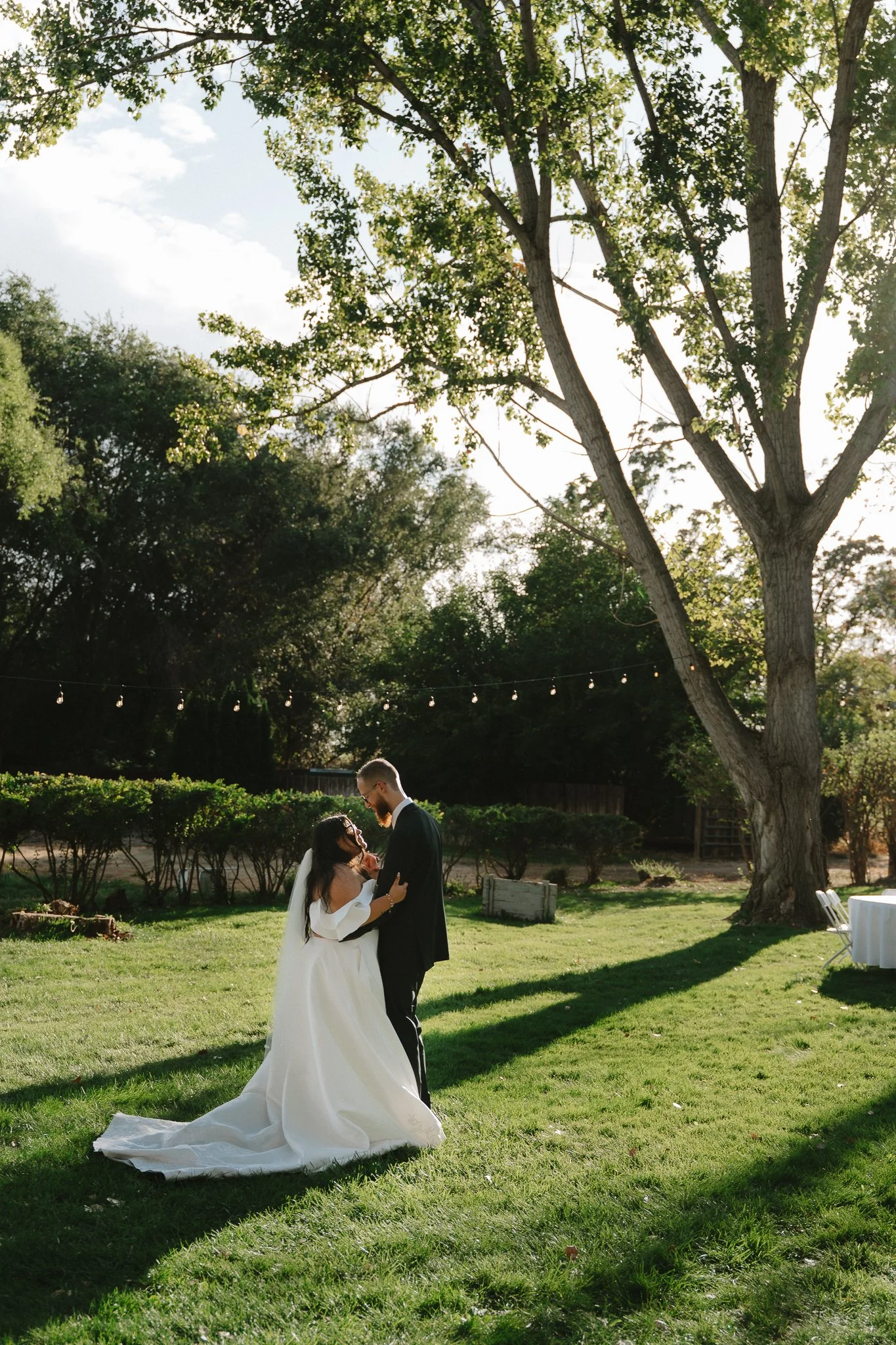 A bride and groom dance outdoors under a large tree on their wedding day, with sunlight filtering through the leaves.