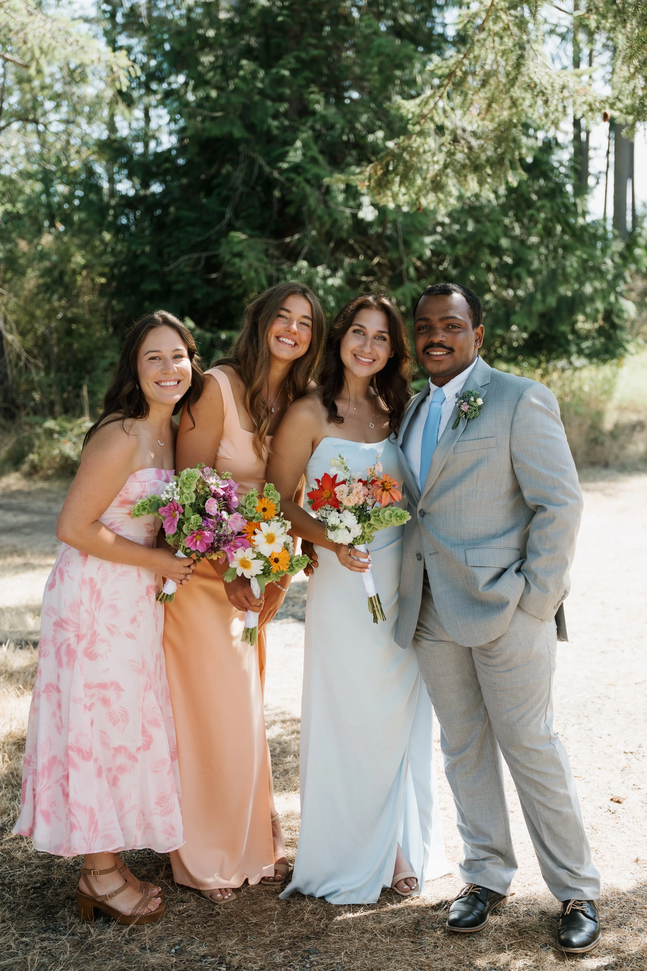A group of four women and one man standing outdoors in a wooded area, dressed in formal attire, with three women holding bouquets of flowers, during a wedding celebration.