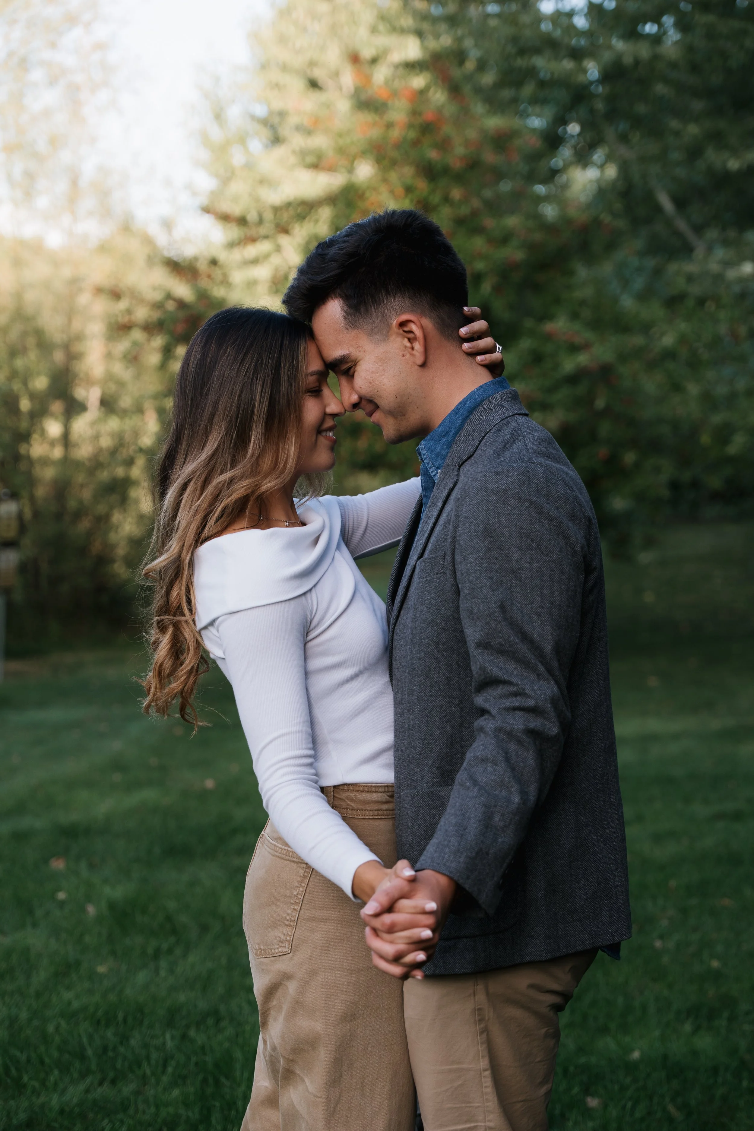 A young couple with their foreheads touching, smiling, and holding hands outdoors in a park setting with green grass and trees in the background.