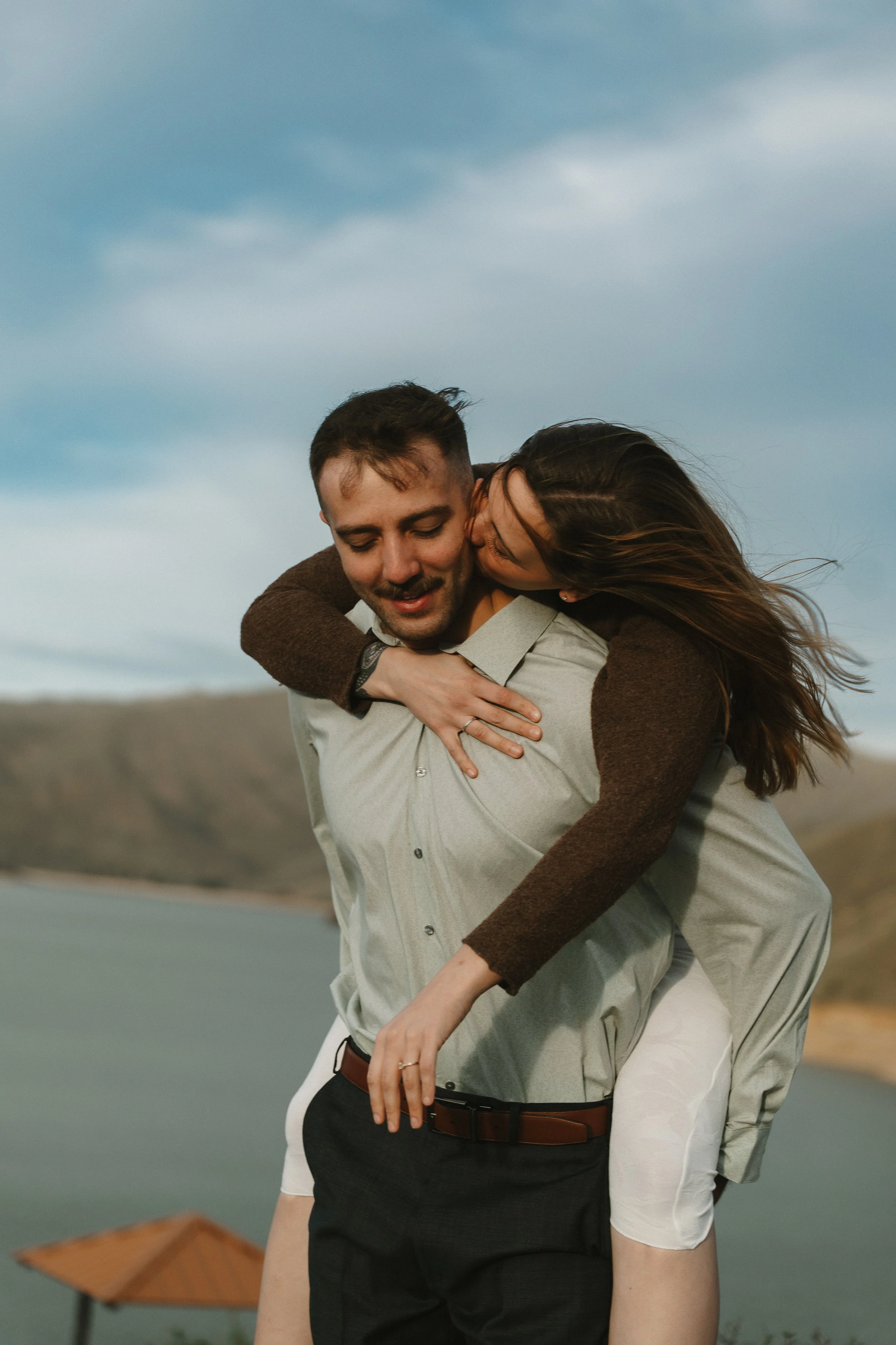 A couple enjoying a piggyback ride outdoors near a body of water, with hills in the background and a cloudy sky.